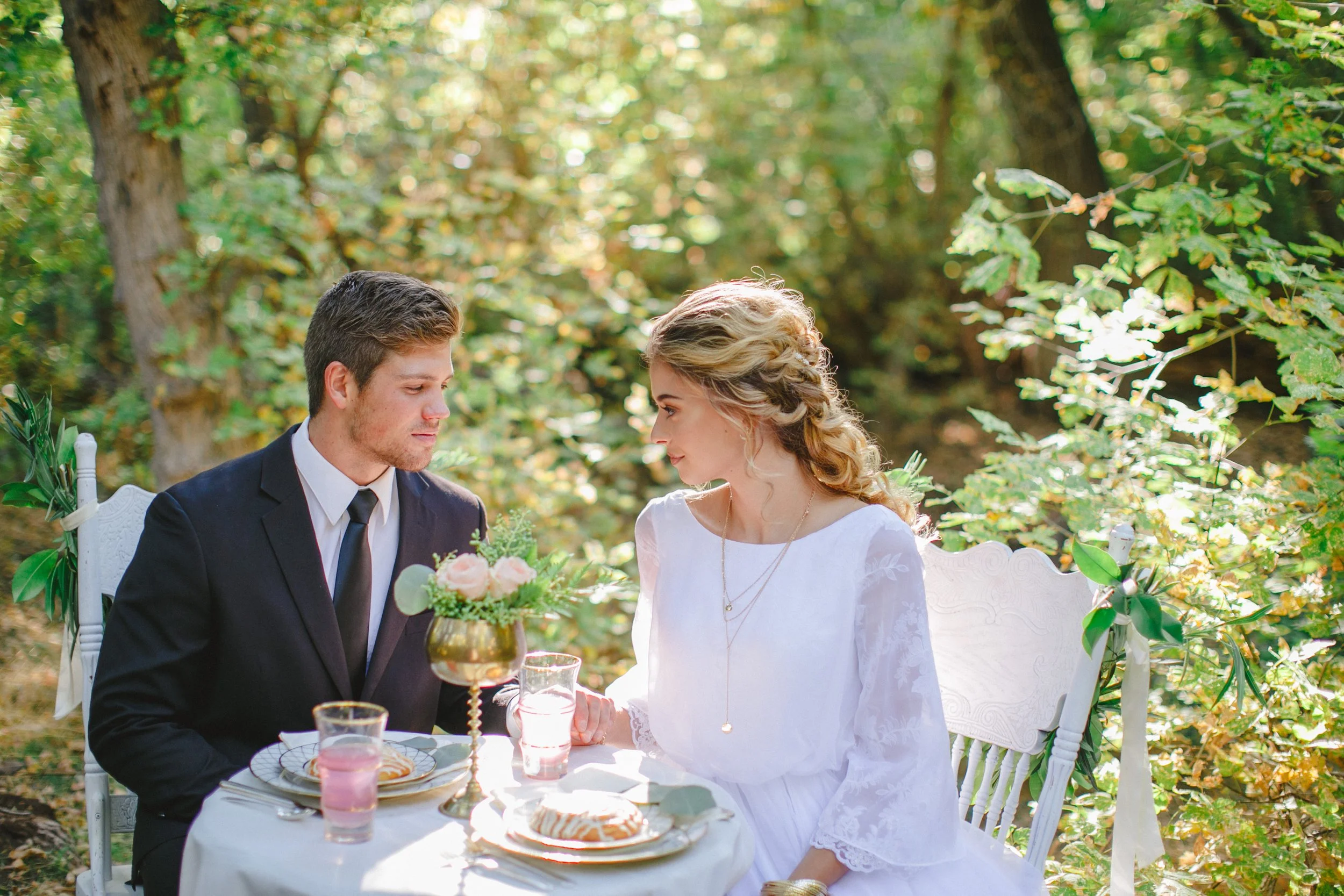 A man and woman sitting at a table outdoors during a wedding or formal event, exchanging eye contact. The man wears a dark suit and white shirt, while the woman is dressed in a white gown. The table has desserts and drinks, and there are trees with g