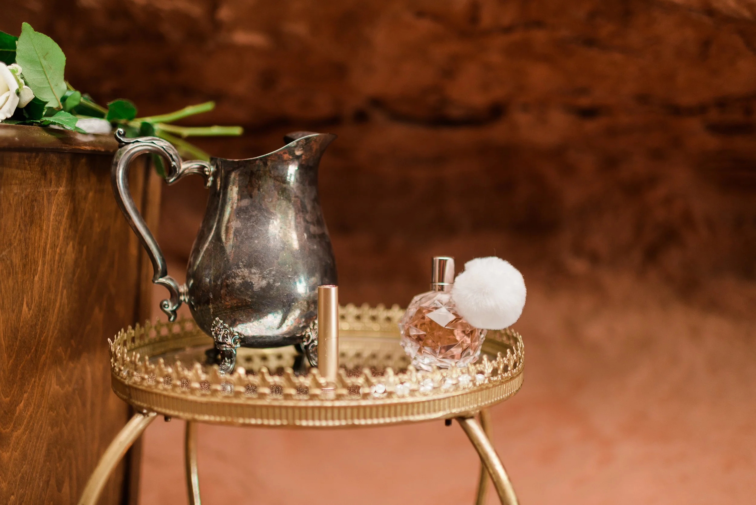 A silver vintage pitcher on a decorative gold tray, with a small perfume bottle, a white fluffy puff, and a gold-capped roll-on perfume bottle, all placed on a round gold table with a wicker edge, against a wood-paneled background.