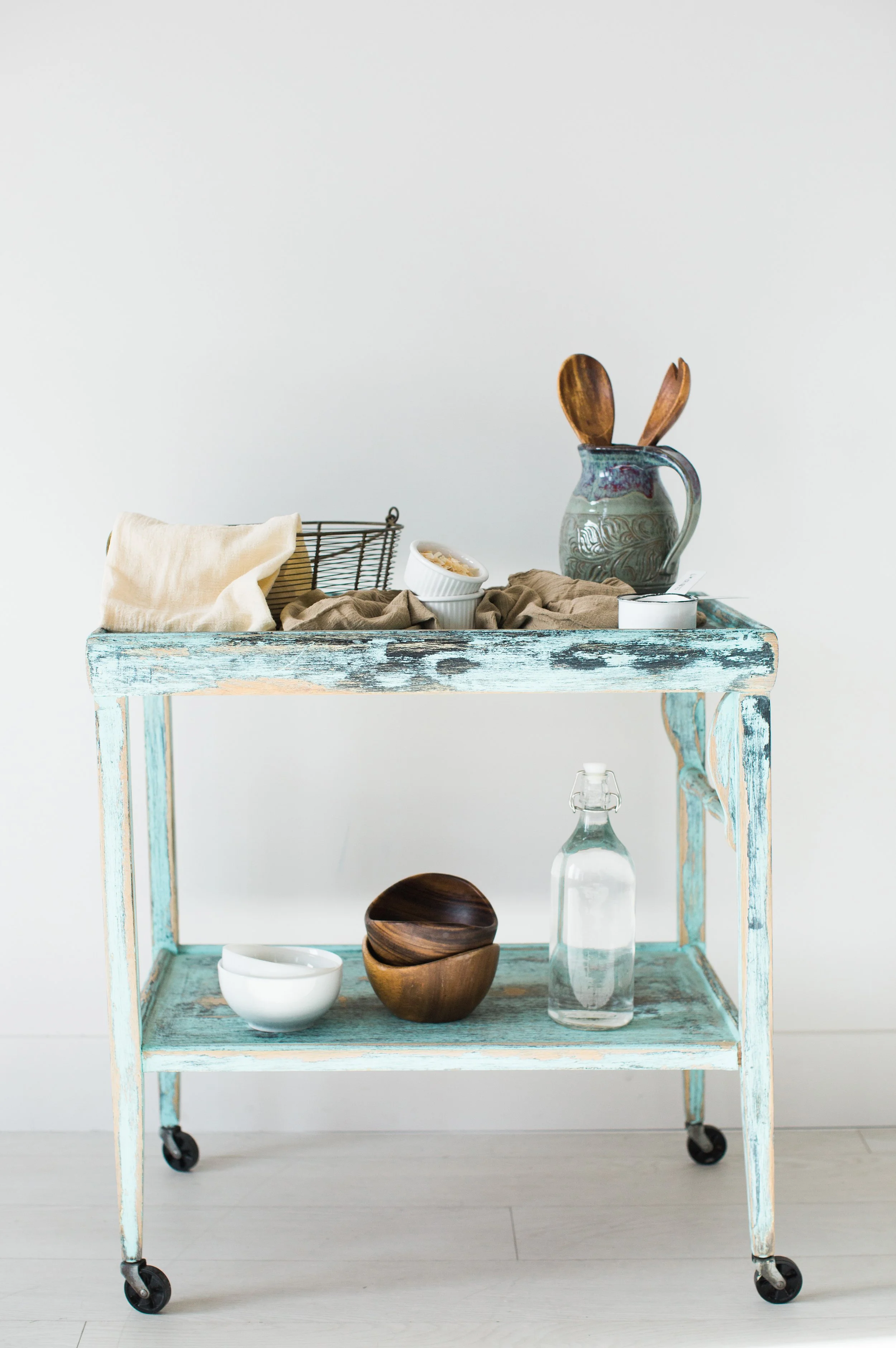 A vintage wooden cart with blue paint, wheels, and a lower shelf, against a plain white wall. The cart holds a ceramic pitcher with wooden utensils, a bowl, a beige cloth, and a wire basket with a cloth inside. The lower shelf contains a small bowl, a wooden container, and a glass bottle.