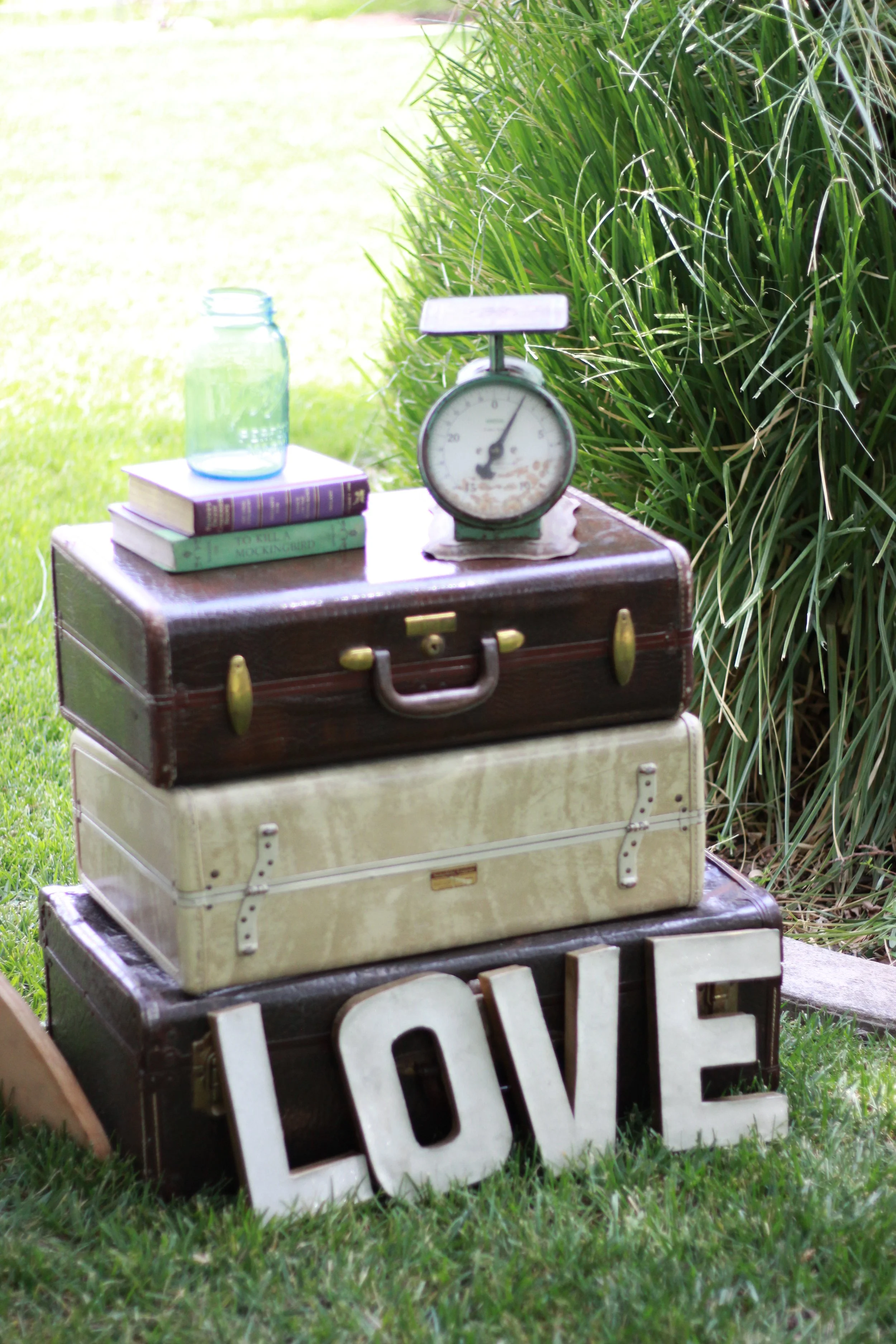 Stacked vintage suitcases with a scale, books, a glass jar, and decorative signs spelling 'LOVE' on grass, outdoors.