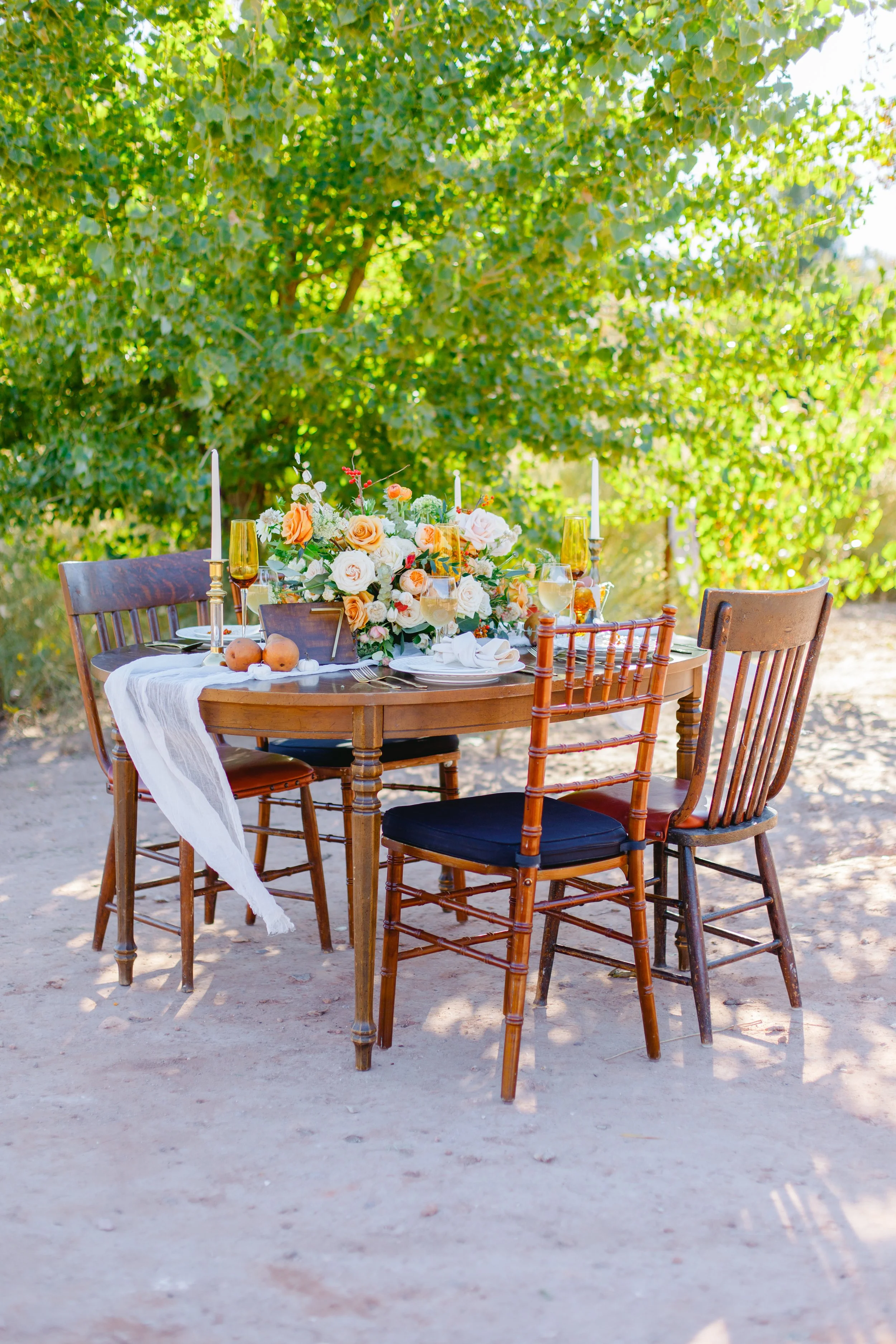 Outdoor dining table set with a floral centerpiece, candles, and drinks, surrounded by chairs, on a dirt surface with green trees in the background.
