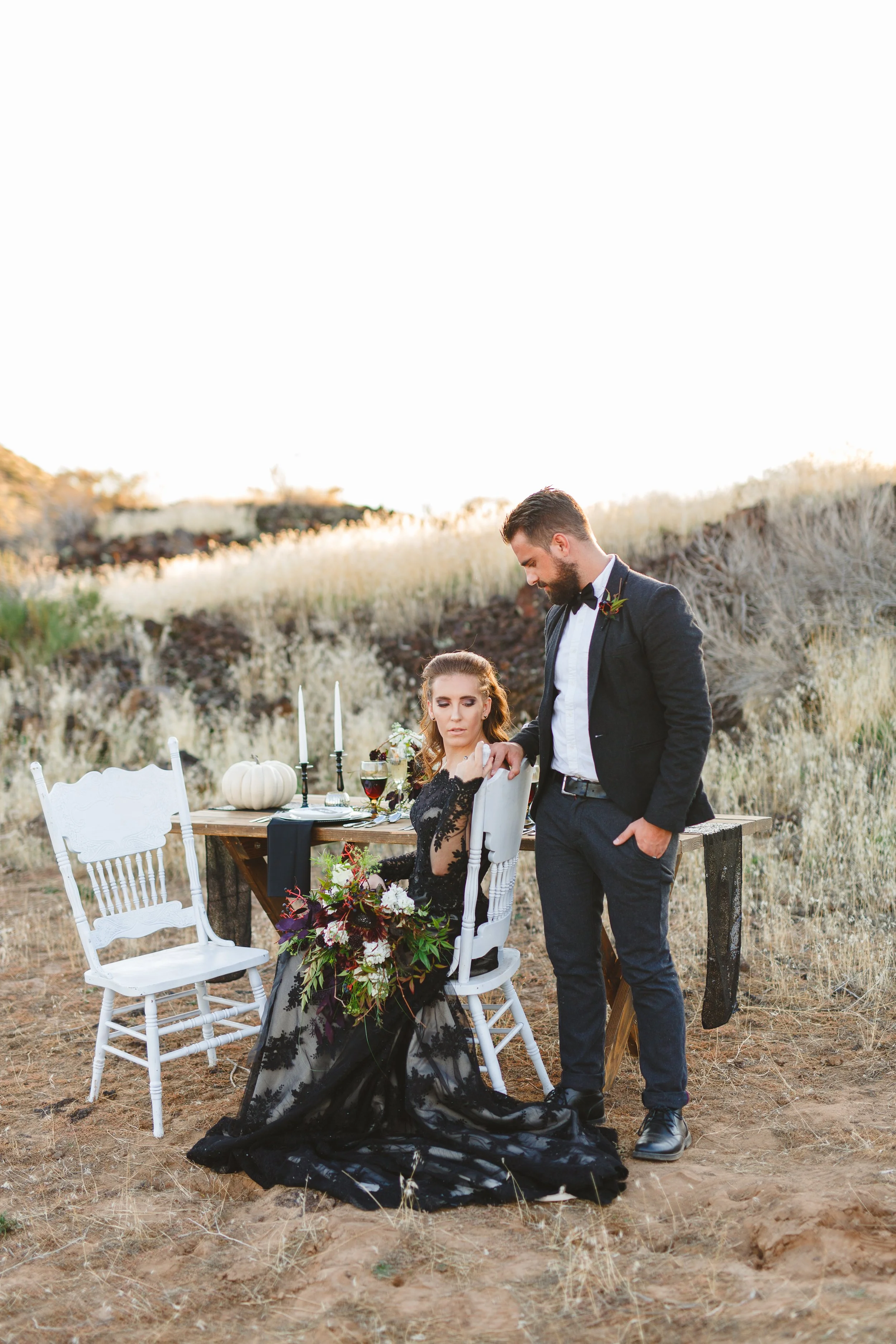 A woman in a black lace gown with floral accents sits at a dining table outdoors in a field, holding a bouquet of flowers. A man in a tuxedo stands beside her, slightly leaning over. The table is decorated with candles, pumpkins, and wine glasses, set against a rustic, natural landscape with dry grasses and rolling hills.