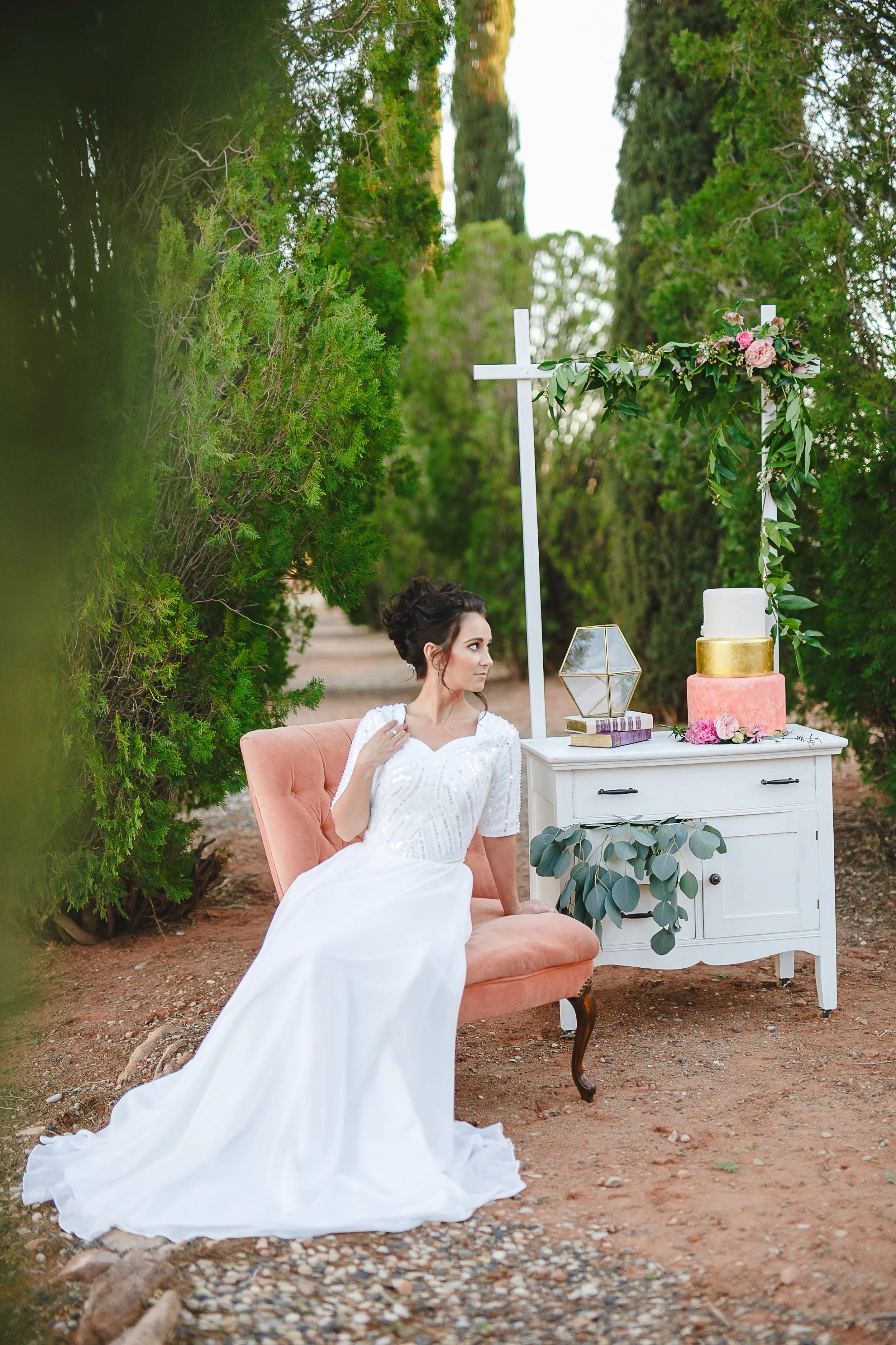 A bride sitting on a peach-colored vintage armchair outdoors next to a white cabinet decorated with a floral arrangement and a wedding cake. The scene is surrounded by green trees and bushes.