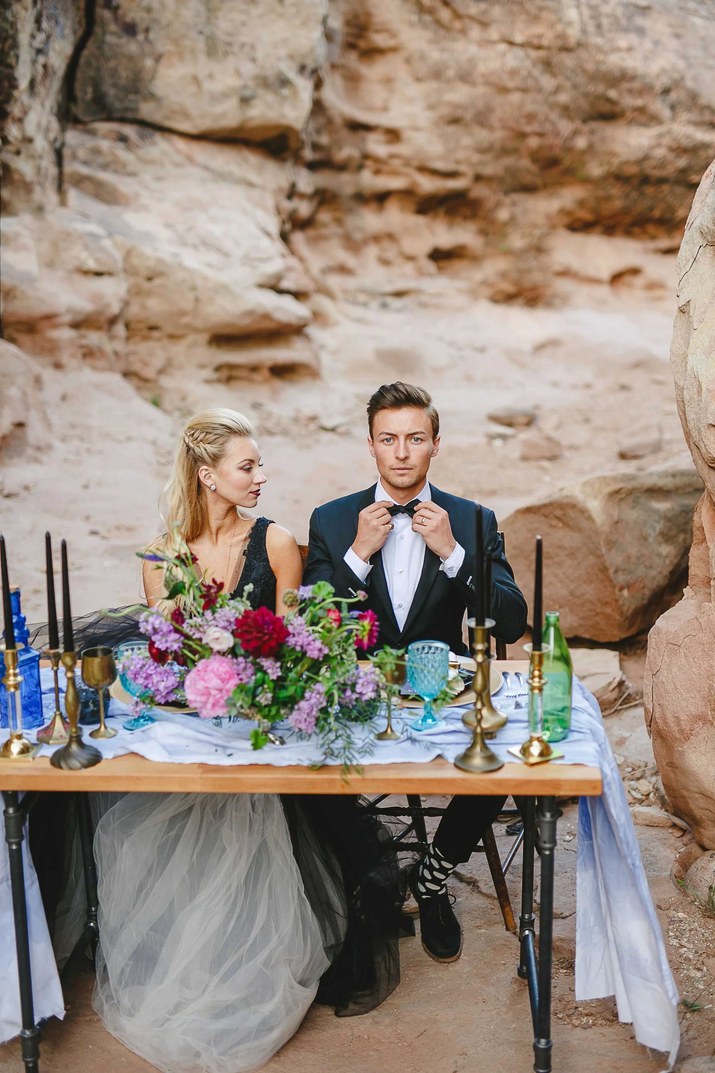 A man and a woman dressed in formal attire sitting at a decorated outdoor table with flowers, candles, and bottles, in a rocky, canyon-like setting.