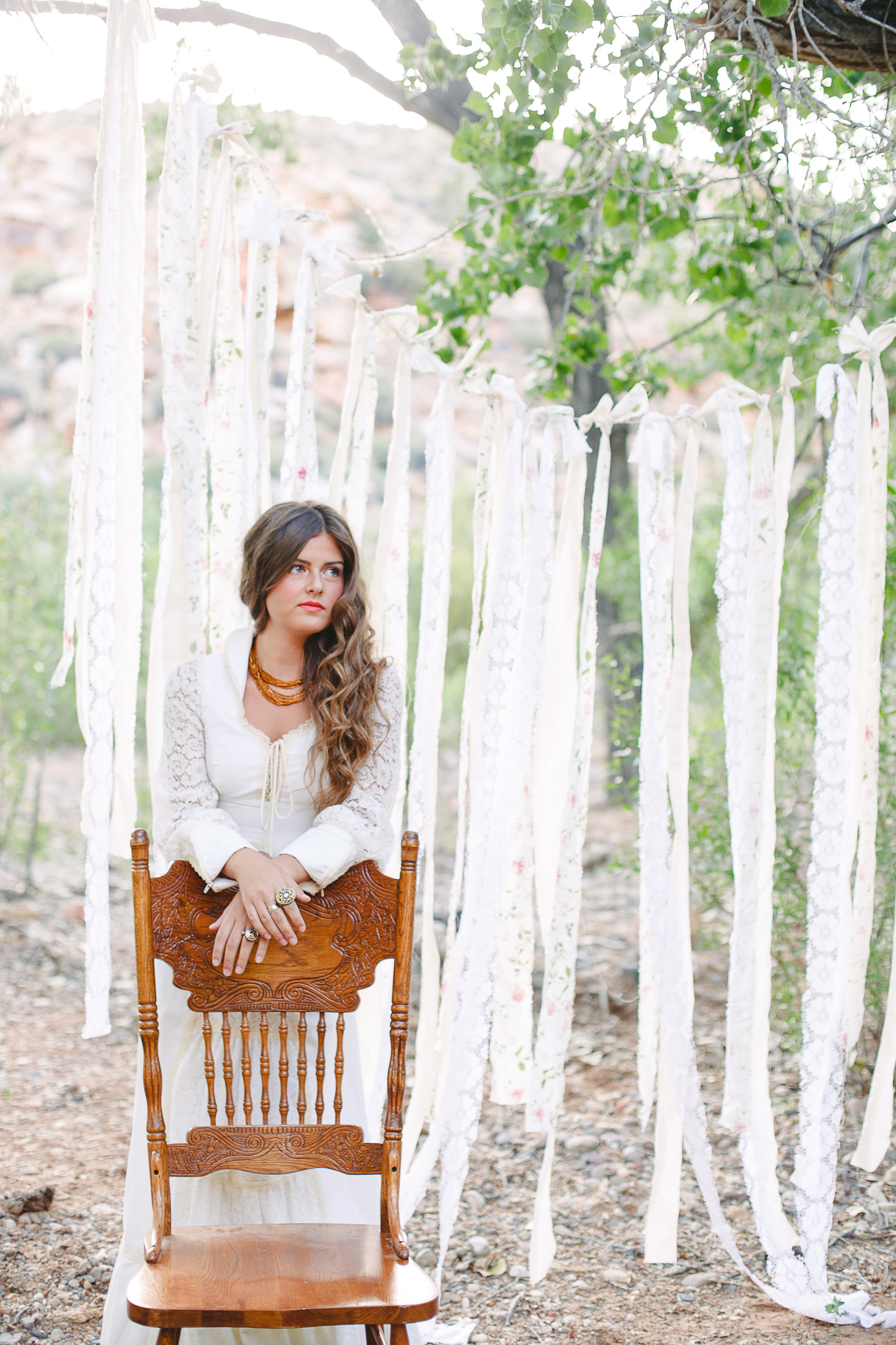 A woman sitting on an ornate wooden chair outdoors with leafy trees and white fabric ribbons hanging in the background.