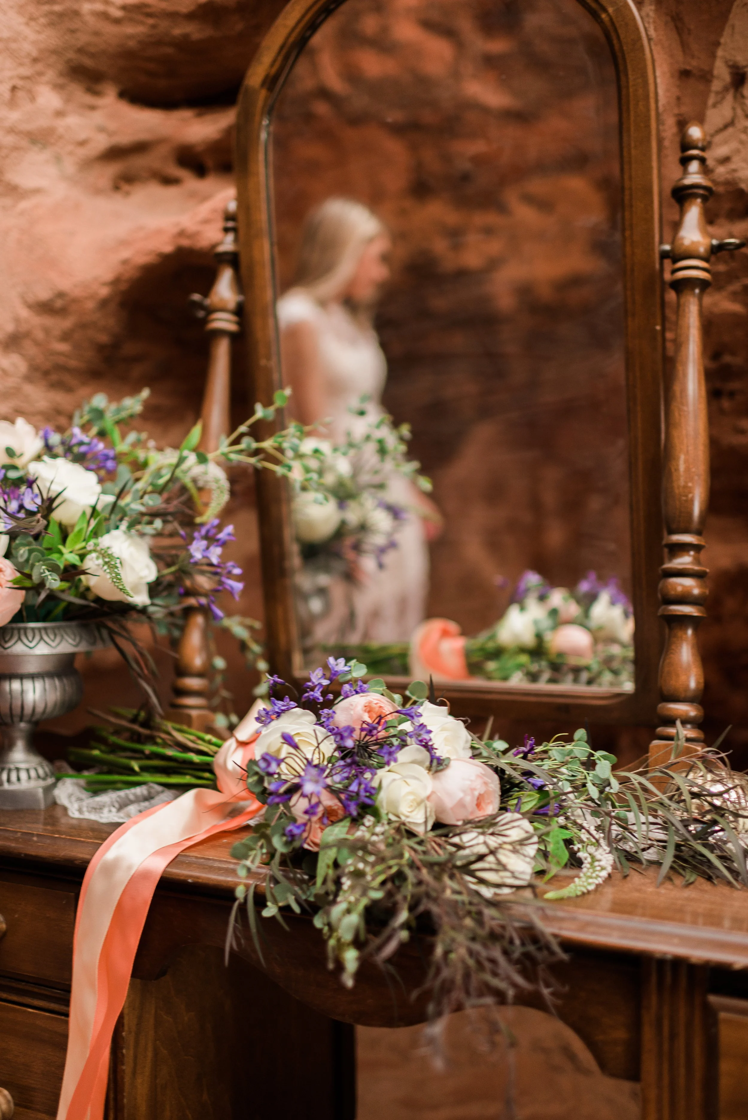 A wooden vanity with a large mirror reflects a woman in a white dress surrounded by bouquets of flowers including roses, lavender, and greenery, resting on the dresser with peach-colored ribbons.