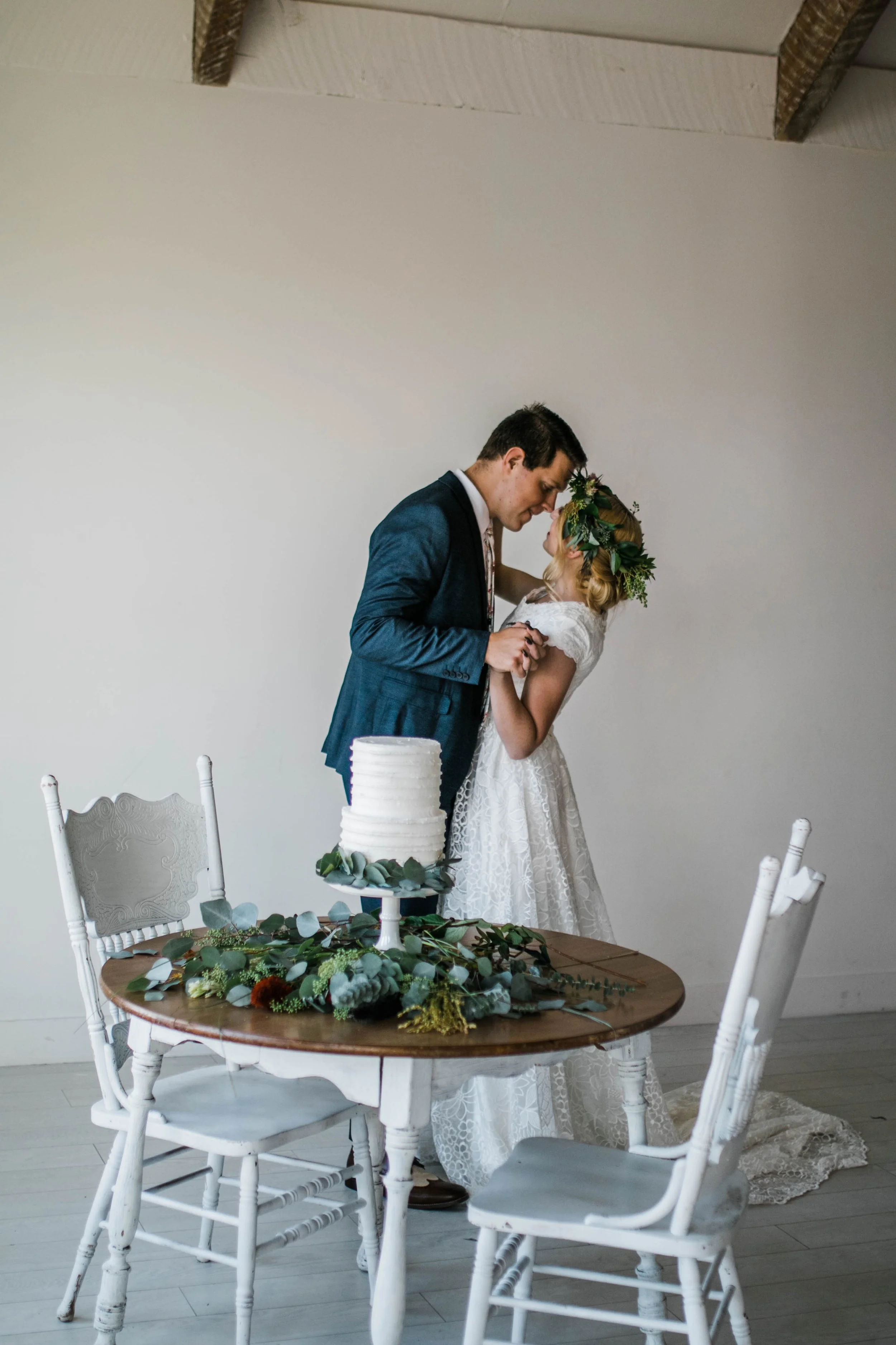 A bride and groom sharing a moment at their wedding reception, leaning their foreheads together. In the foreground, a round table decorated with greenery and a white, tiered wedding cake is visible. The setting has white chairs and a minimalist, light-colored background.