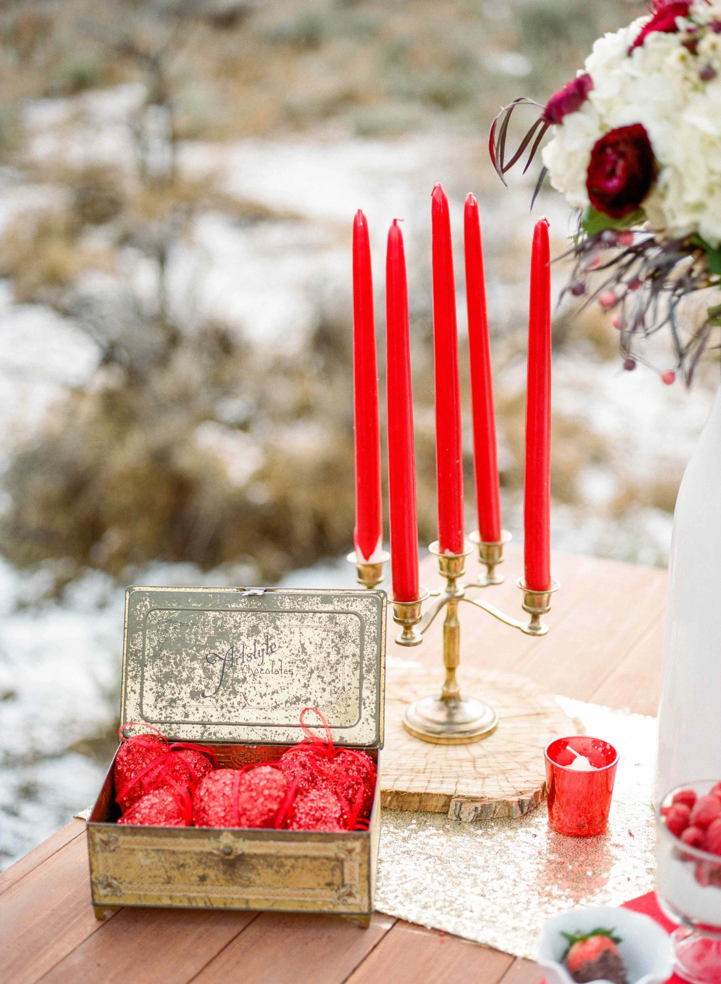 A table with red decorative apples, a vintage-style cookie tin with the words 'Artful Chocolates' on the lid, five tall red candles in a brass candle holder, a small red cup with a white candle, and a blurred background of an outdoor scene with snow and trees.