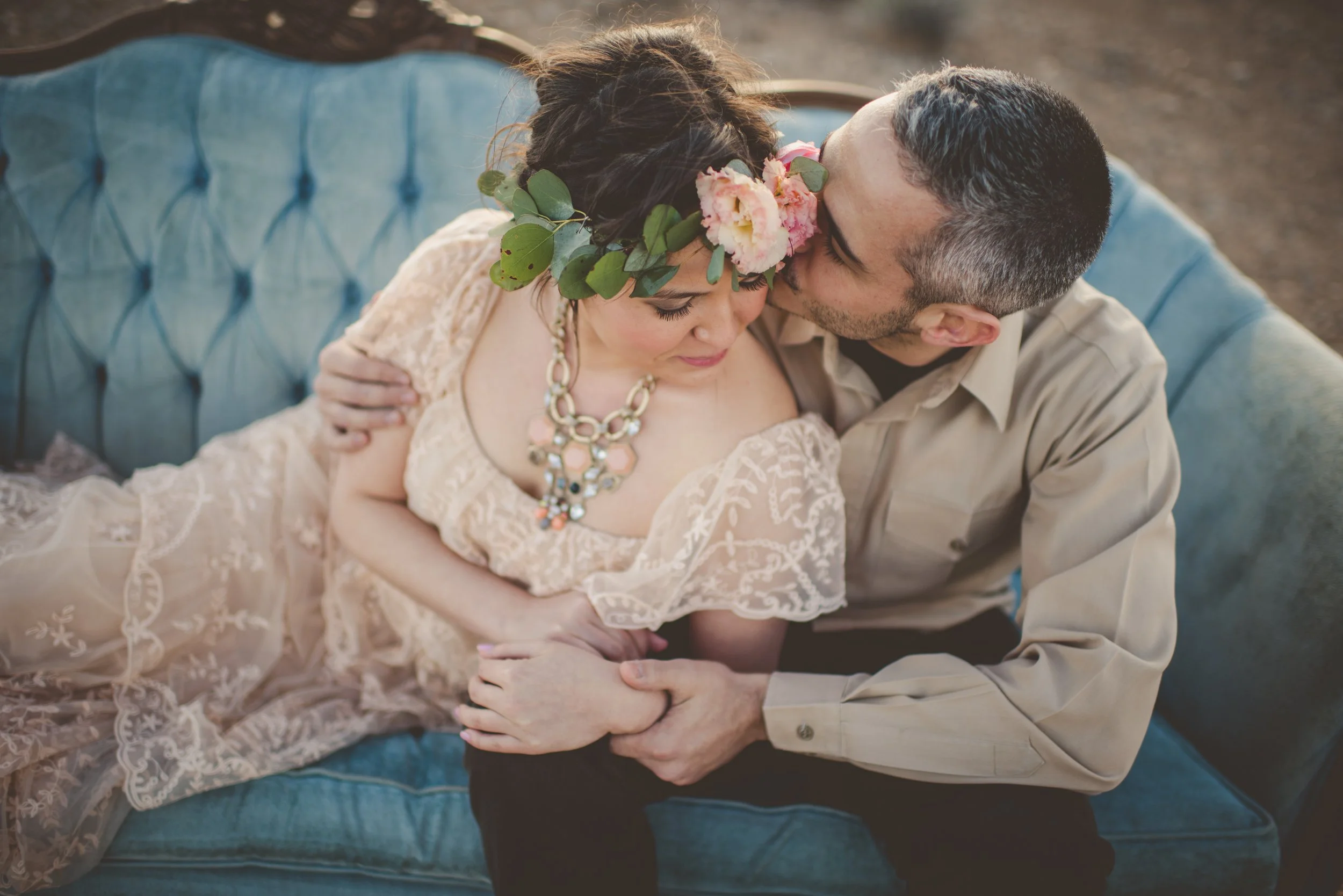 A couple sitting on a blue upholstered vintage sofa, sharing a romantic moment with their foreheads touching. The woman has dark hair styled in loose waves, adorned with a pink flower and green leaves, wearing a lace dress with floral embroidery. The