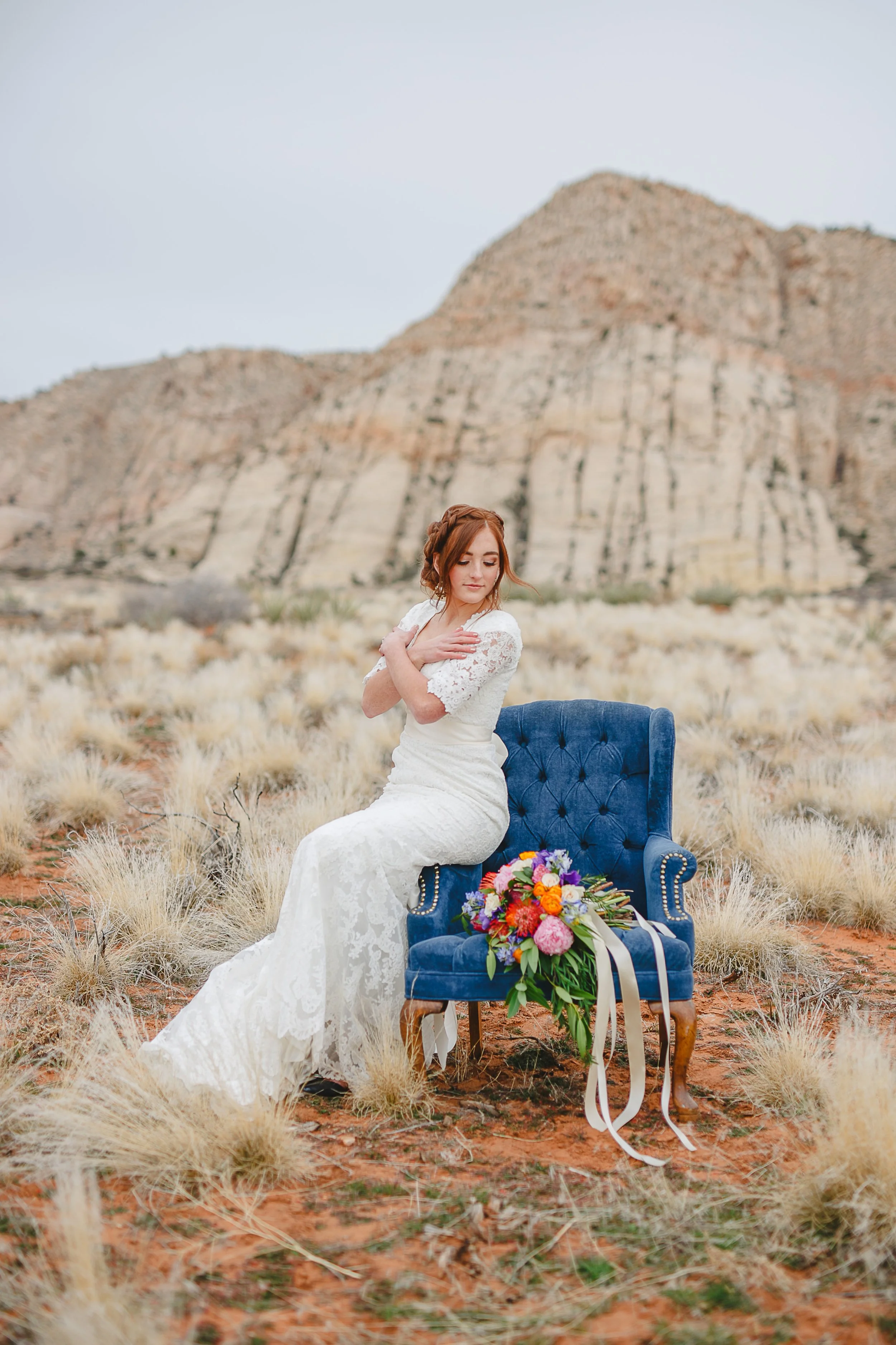 A woman in a white lace wedding dress is sitting on a blue velvet Victorian-style chair in a desert landscape with rocky hills in the background. She's holding her arms crossed over her chest and has a bouquet of colorful flowers resting on the ground.