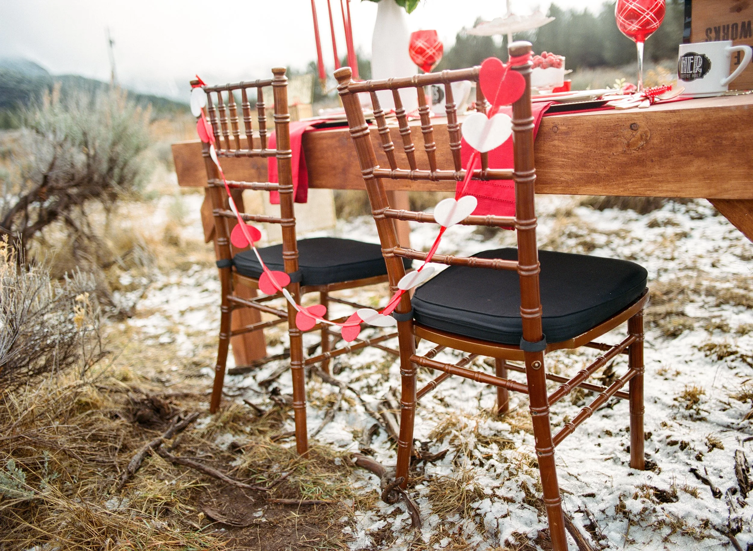 A wooden outdoor table with chairs decorated with red and white paper hearts and red heart and heart-patterned lanterns, set outdoors in a natural setting with grass, dirt, and light snow, trees and hills in the distance.