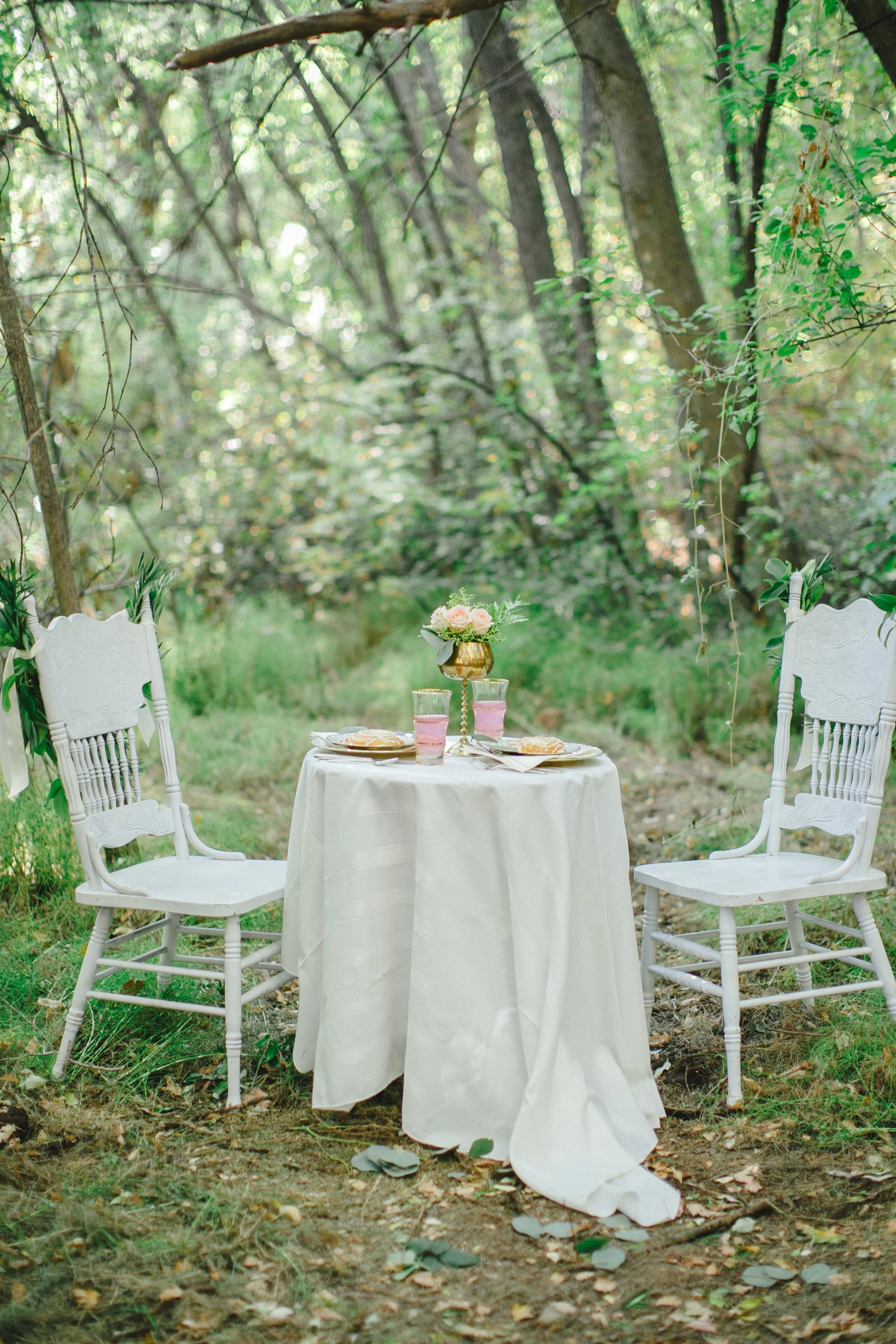 A round table covered with a white cloth set for a meal outdoors in a wooded area. Two white chairs with decorative backs and green ribbons are placed on either side of the table. In the center of the table is a gold vase with pink and white flowers, surrounded by pink glasses and autumn leaves scattered on the ground.