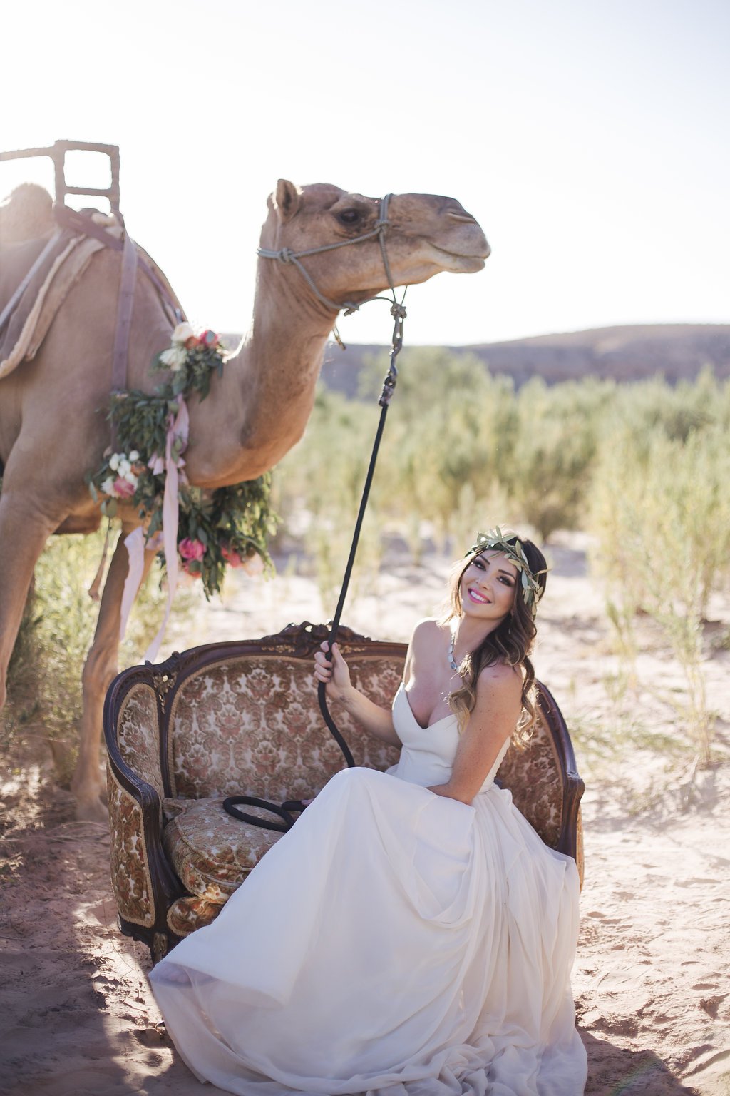 A woman in a white dress sitting on an ornate vintage sofa outdoors, holding the reins of a camel decorated with flowers, in a scenic desert-like landscape with green trees and hills in the background.