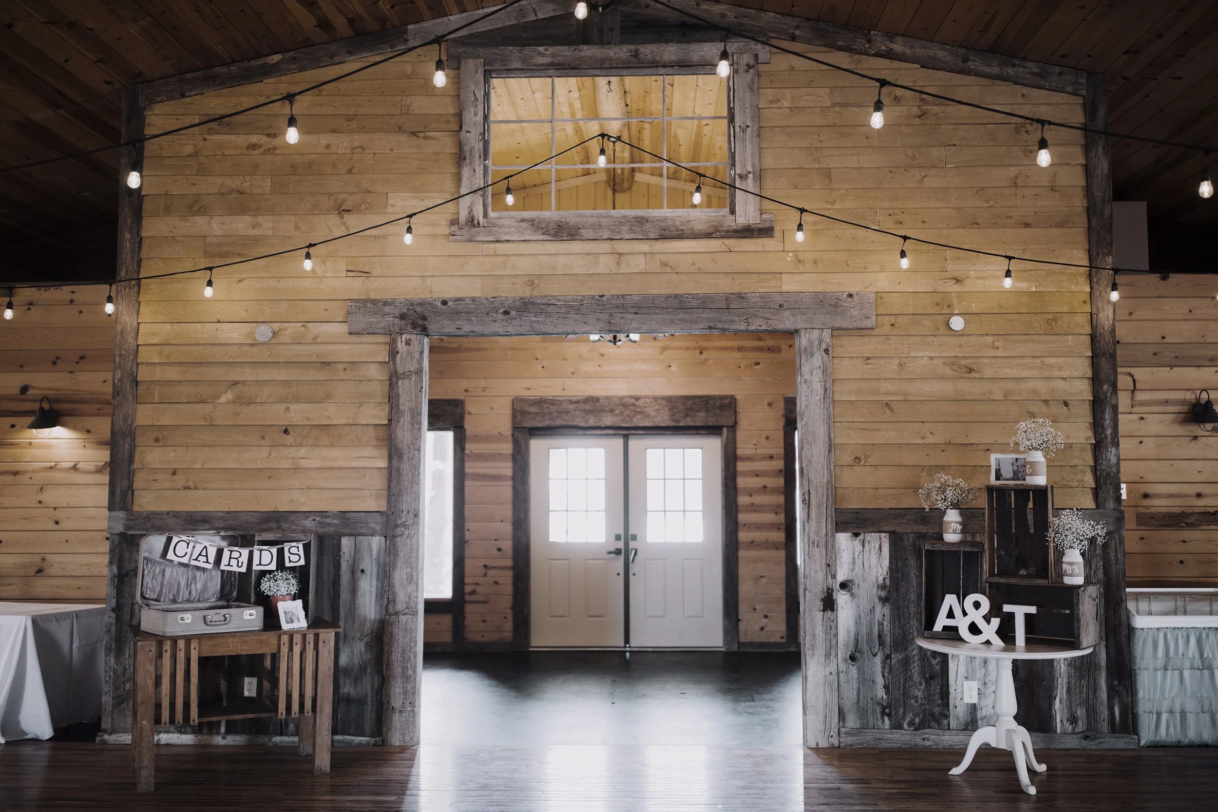 Rustic indoor wedding or event space with wooden walls and ceiling, string lights hanging across the room, and decorated tables with signs and flowers.