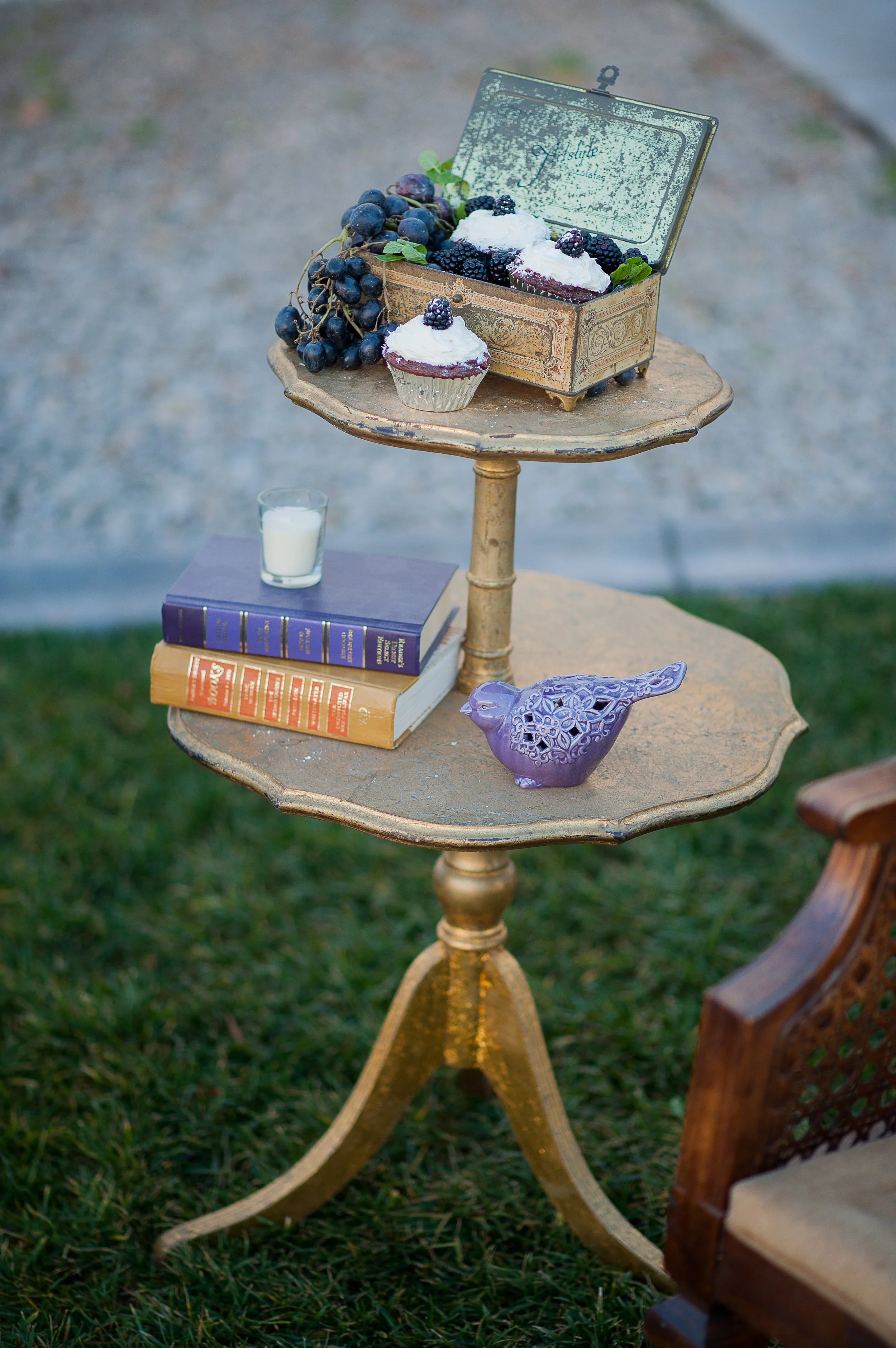 A small vintage table with a gold finish on grass, displaying a decorative box filled with blackberries and grapes, a cupcake with blackberries on top, a glass of milk, and stacked books. There is an ornate purple bird ornament on the table.
