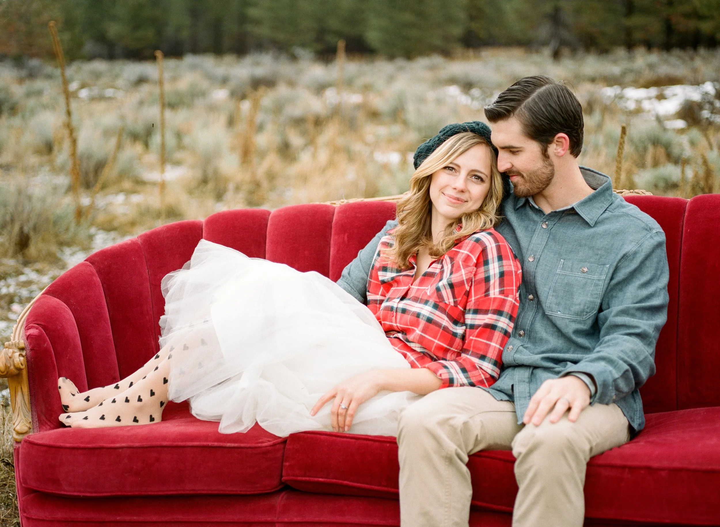 A young couple sitting closely together on a red velvet sofa outdoors in a natural setting, with the woman smiling and the man leaning in affectionately.