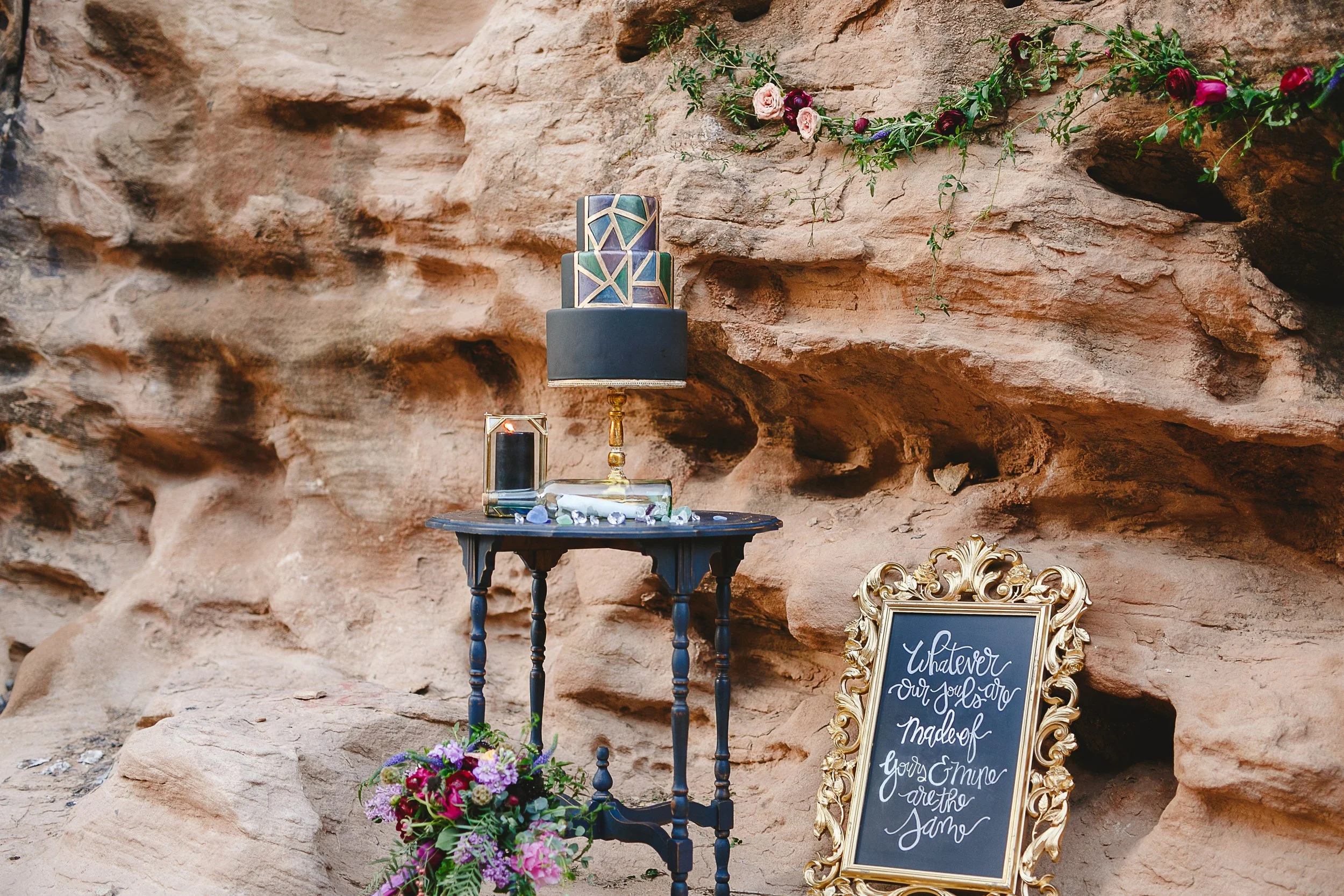 Decorative setup against a rocky background featuring a black small table with a colorful patterned candle holder and a candle. A floral bouquet and a blackboard with elegant writing are also present, indicating a celebration or special event.