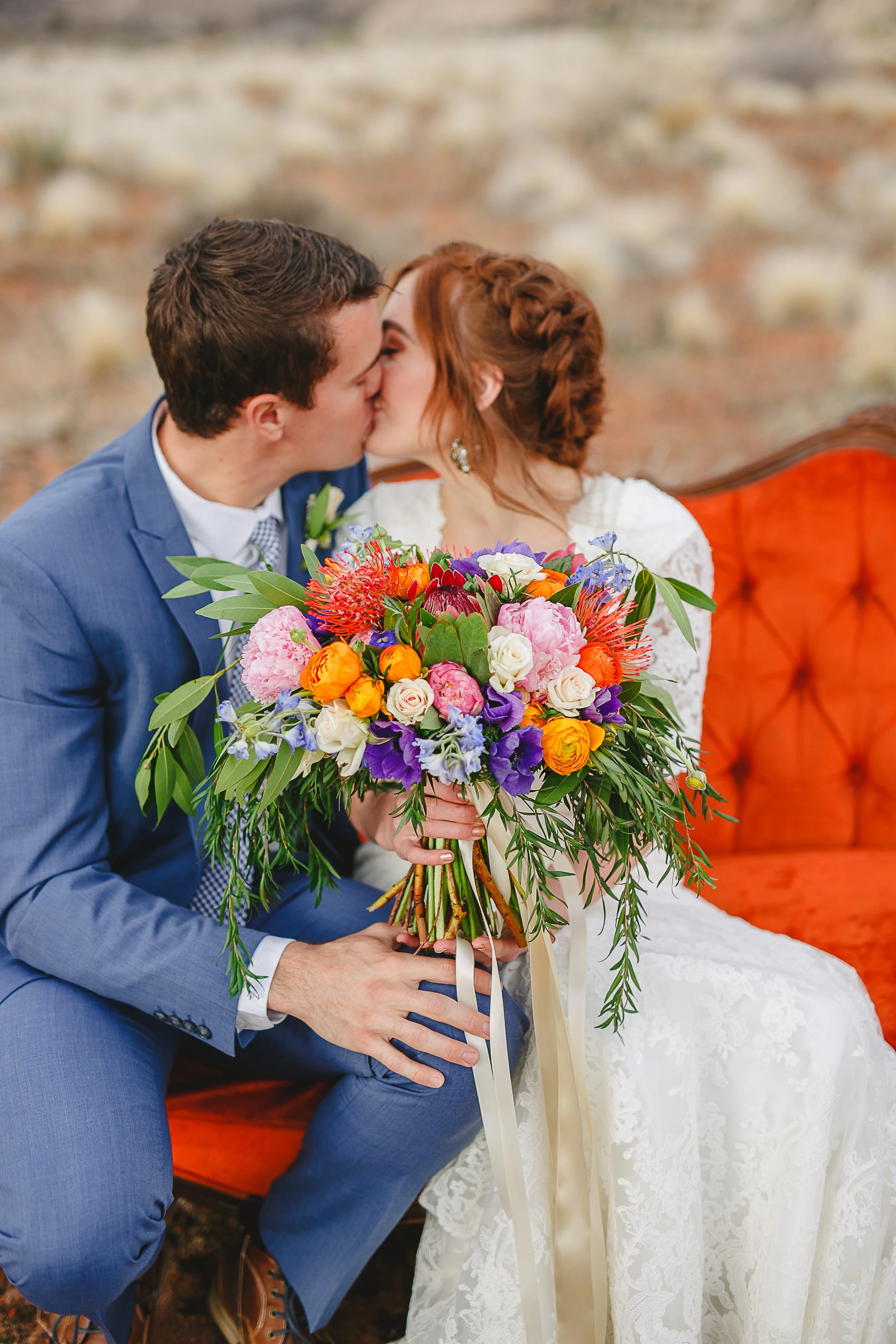 A couple sharing a kiss during their wedding, with the groom in a blue suit and the bride in a white lace wedding dress, holding a large bouquet of various colorful flowers.