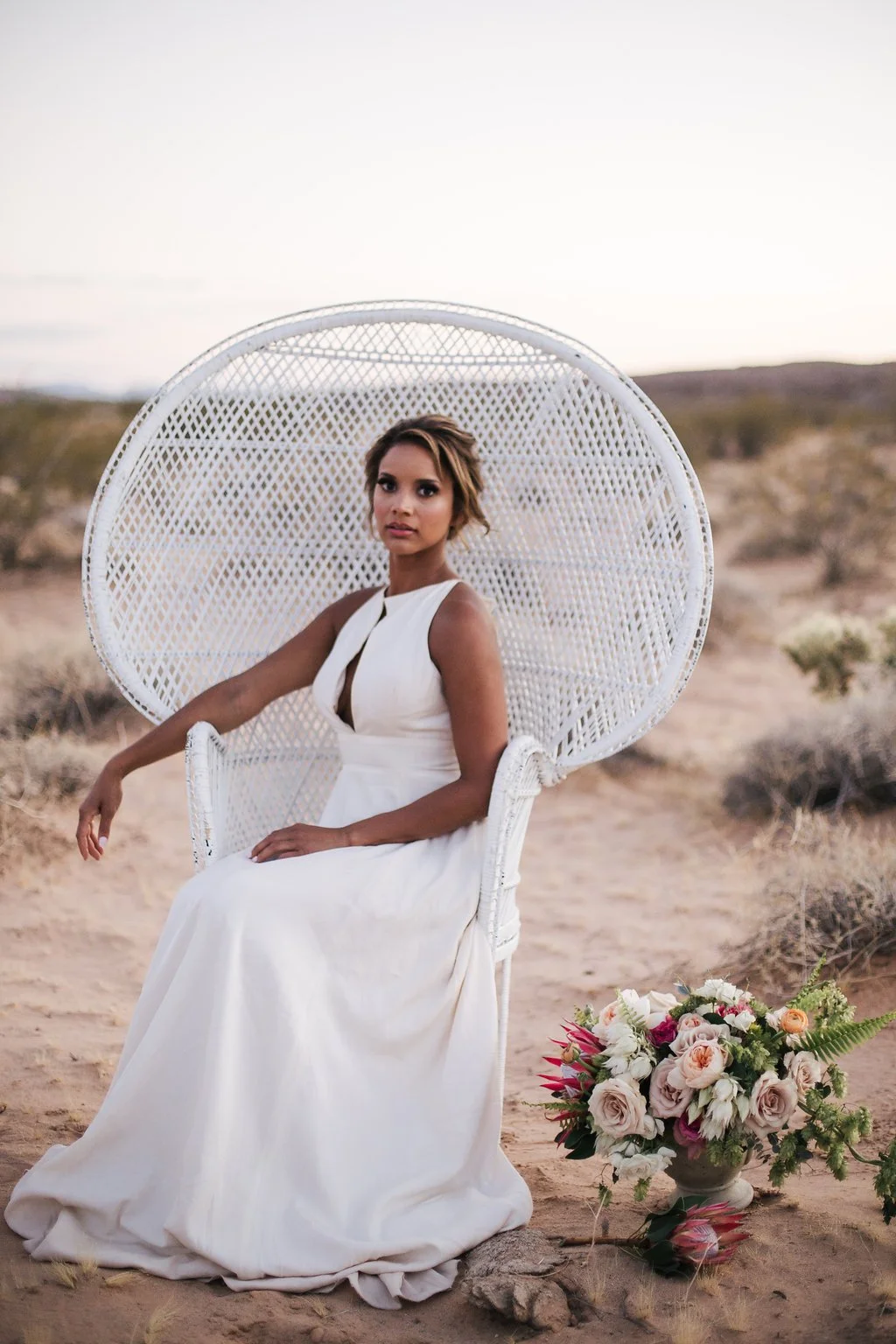 A woman in a white dress sitting on a white wicker chair in a desert landscape with a bouquet of flowers nearby.