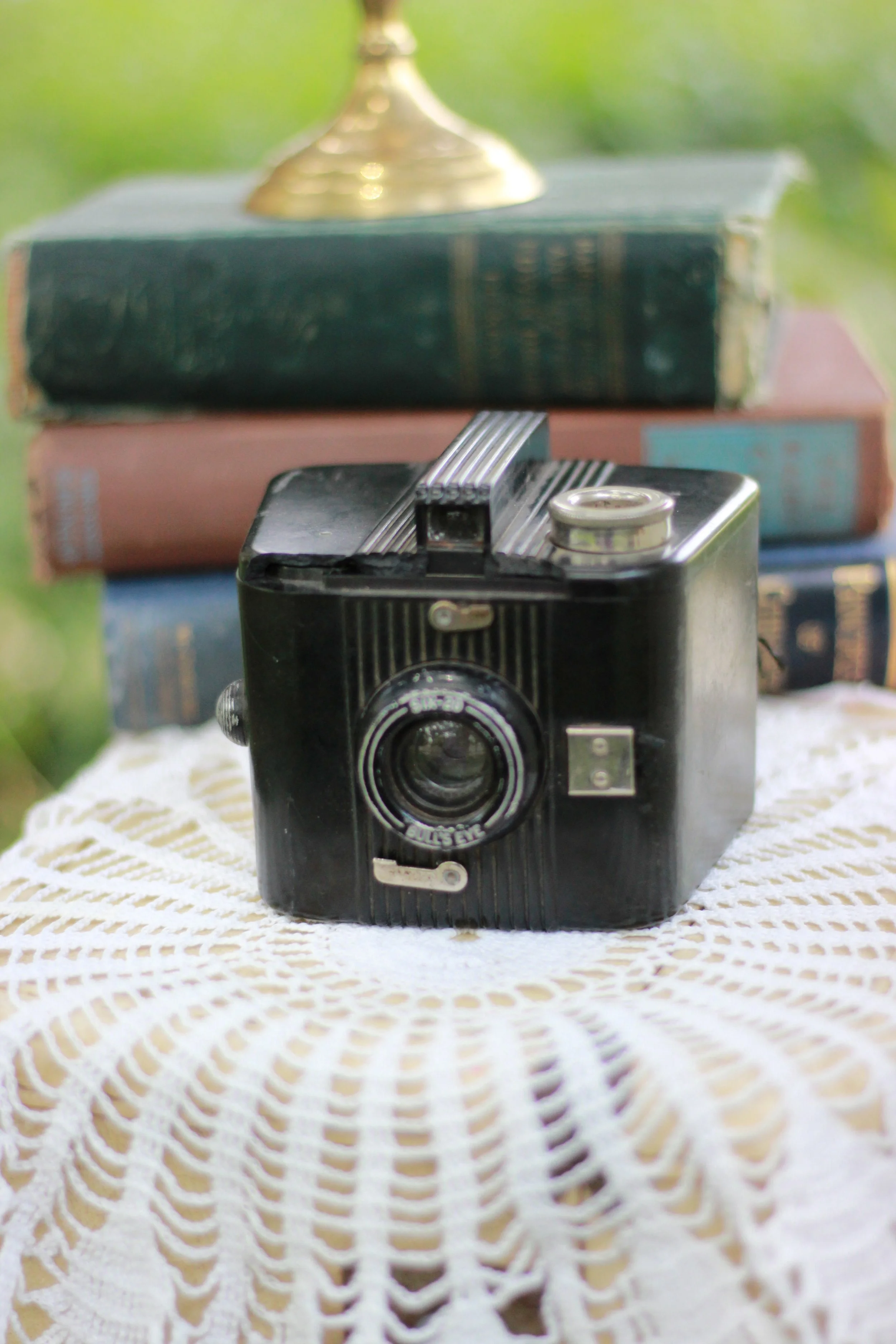 A vintage camera sitting on a white lace tablecloth with a stack of old books and a brass lamp in the background, outdoors with a blurred green background.