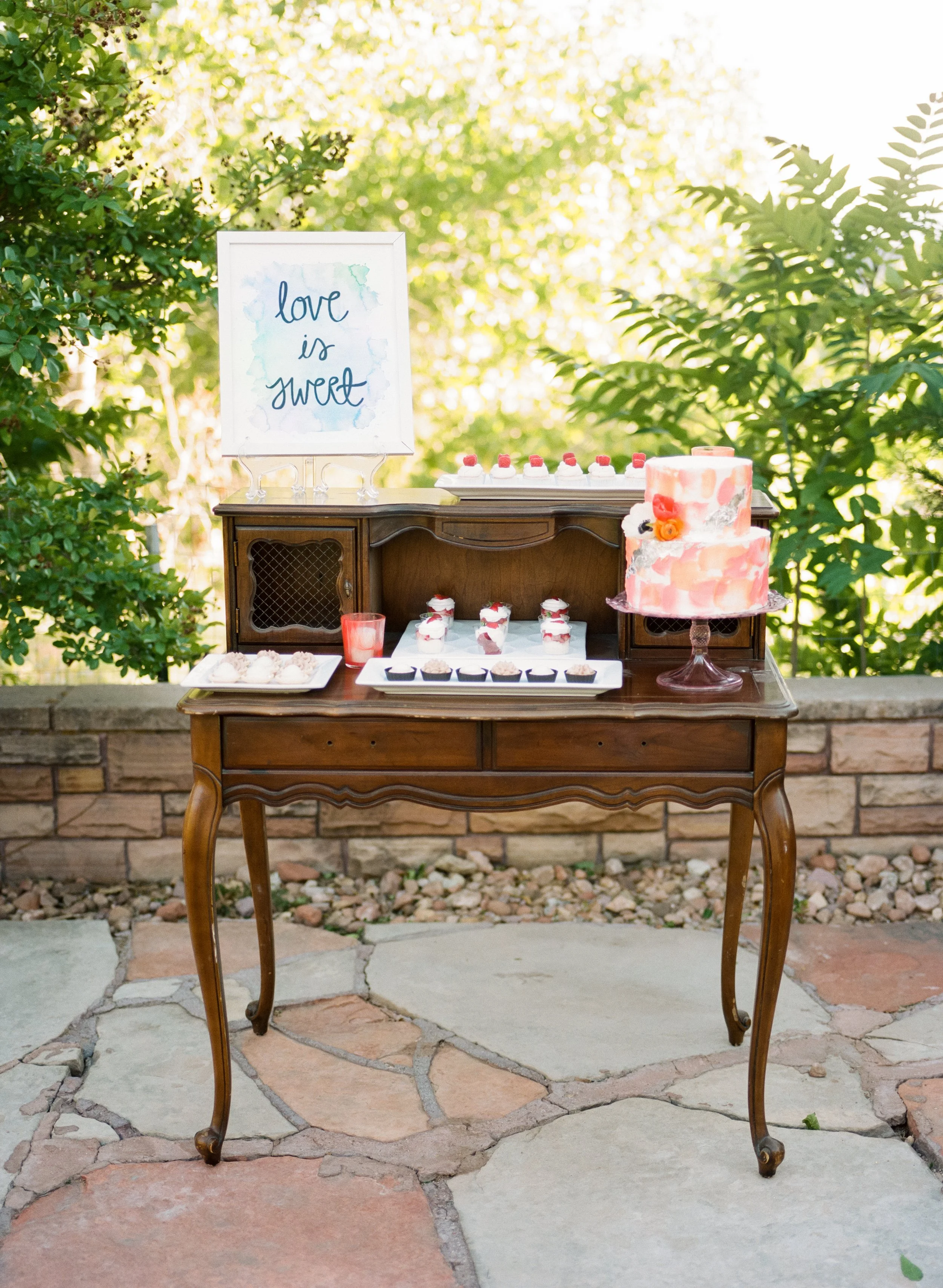 Outdoor dessert table with a watercolor pink and white cake, small cupcakes, and a sign reading 'love is sweet' on a wooden table, surrounded by lush green trees and plants.