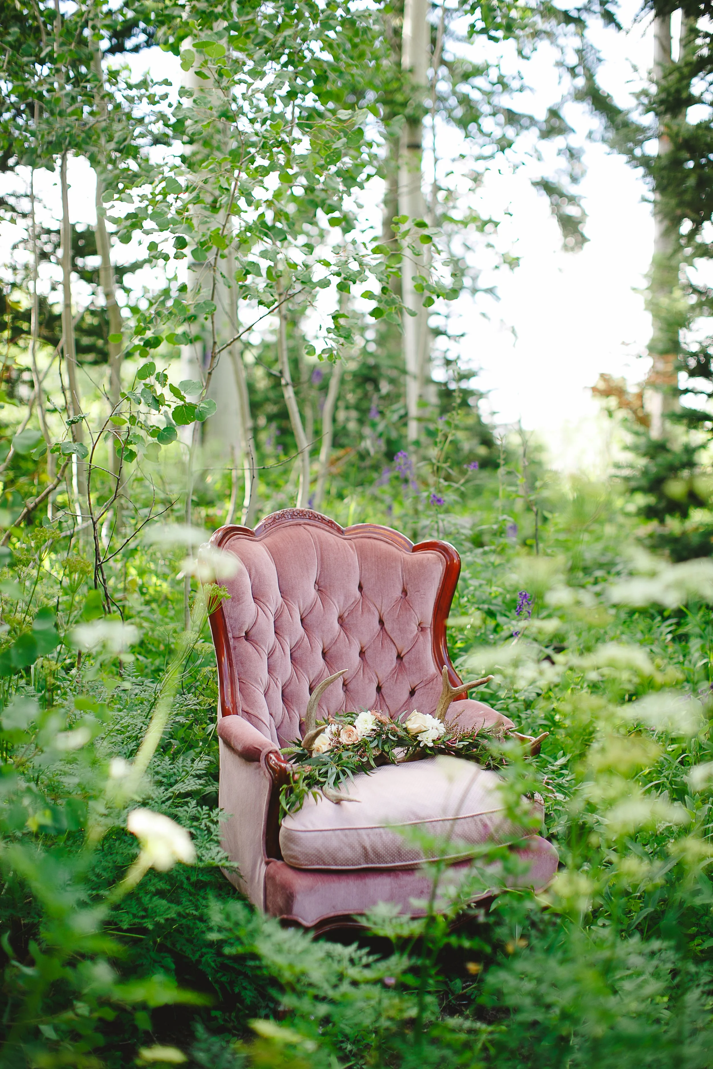 A vintage pink tufted armchair with a floral and antler decoration on the seat, set in a lush green forest with trees and sunlight filtering through the leaves.