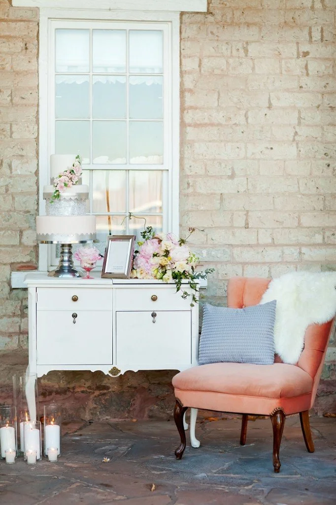 A cozy indoor scene featuring a pink upholstered armchair with a blue pillow and white furry blanket, a white sideboard decorated with pink flowers, a framed picture, and a tiered cake, in front of a brick wall and window with candles on the floor.