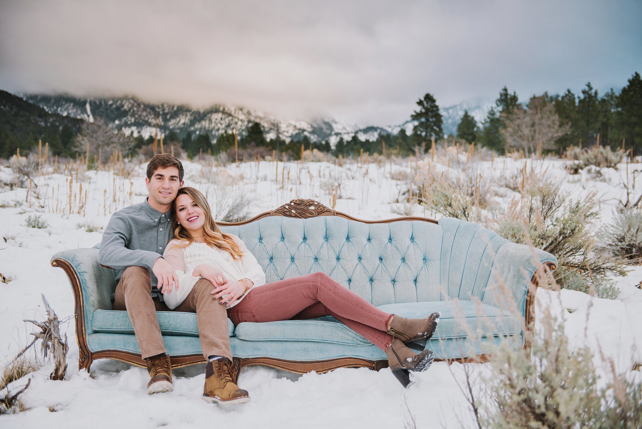 A couple sitting on a vintage blue sofa outdoors on snow-covered ground with mountains and trees in the background.