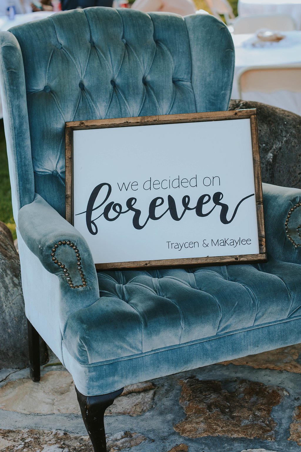A blue velvet armchair with a framed sign resting against the back. The sign says 'we decided on forever' with the names Traycen & Makaylee at the bottom, indicating a wedding or engagement celebration. The chair is on a stone surface outdoors.