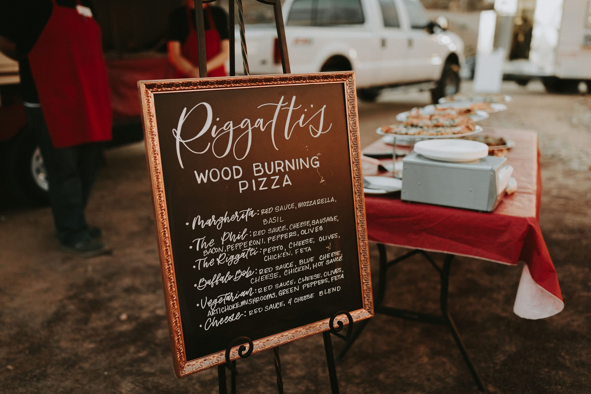 A chalkboard sign advertising wood burning pizza at an outdoor event, with people and vehicles visible in the background.