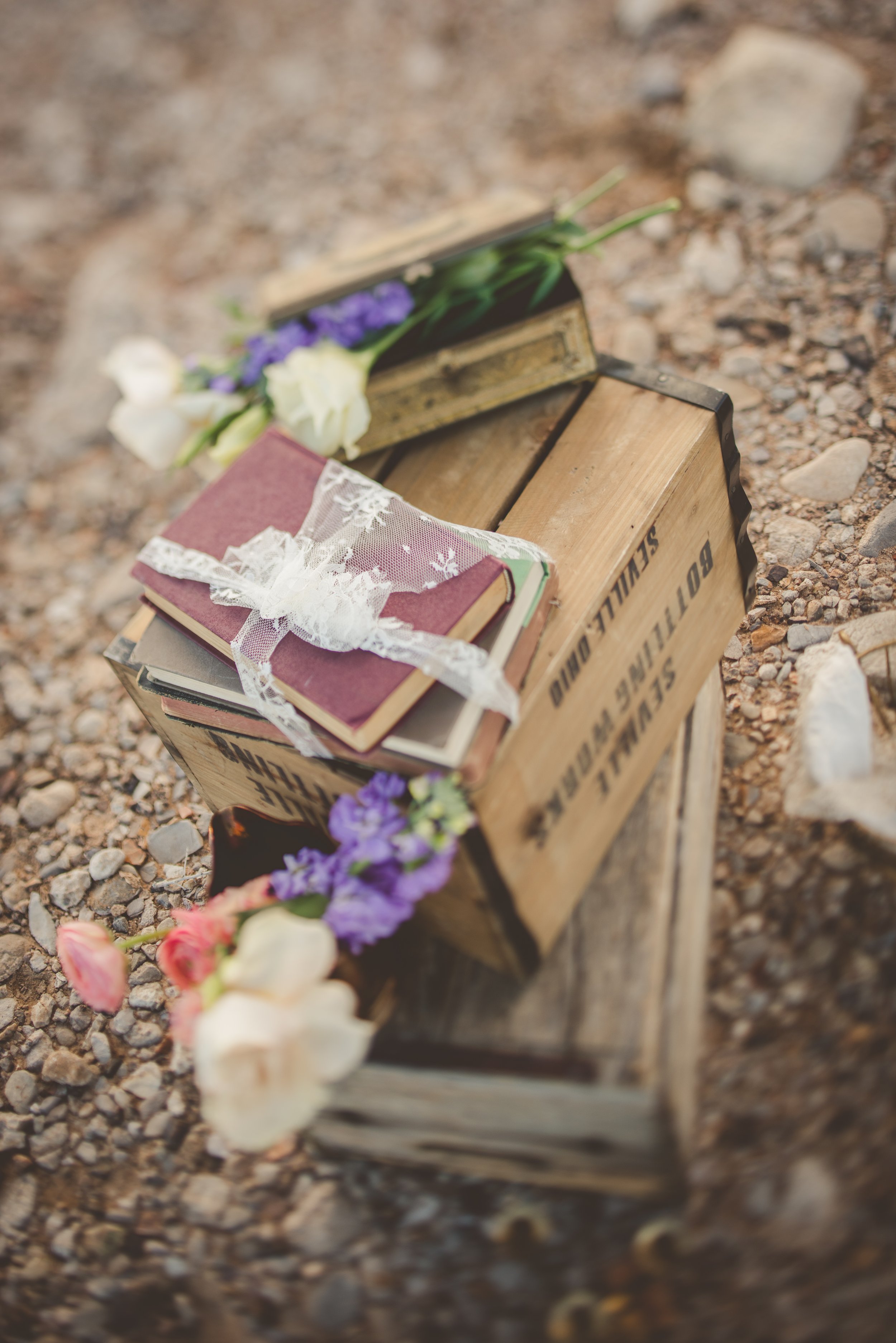 A wooden crate with a vintage look sits on a gravel ground. It contains closed books with a lace ribbon, and among the books are purple and white flowers, some of which are spilling out onto the ground.