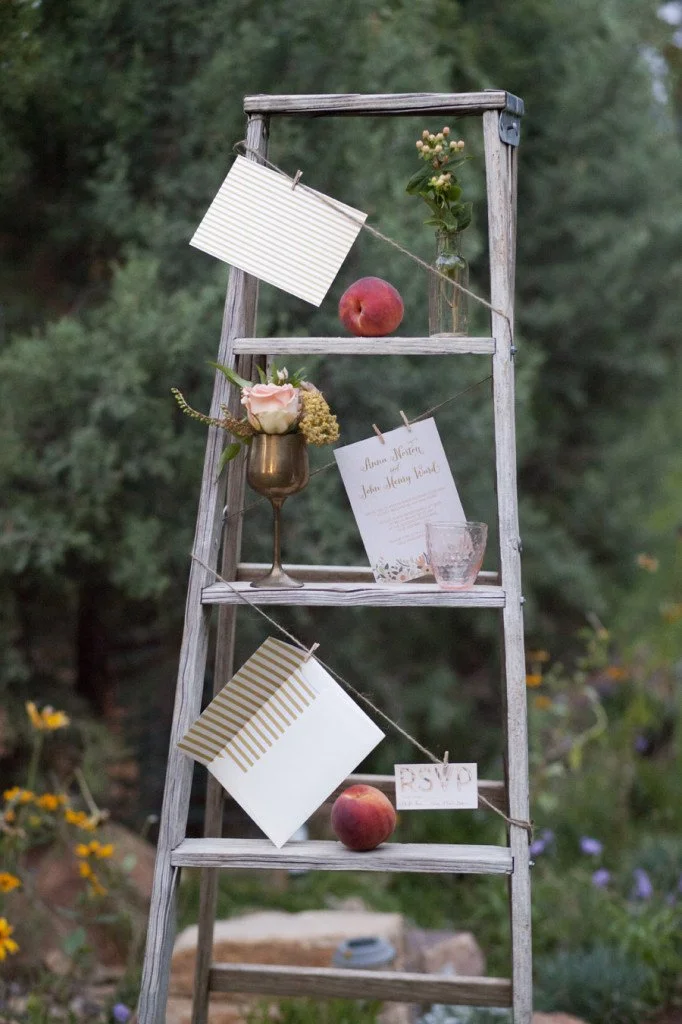 A rustic wooden ladder decorated with flowers, peaches, and paper cards hanging from strings, set outdoors in a garden.