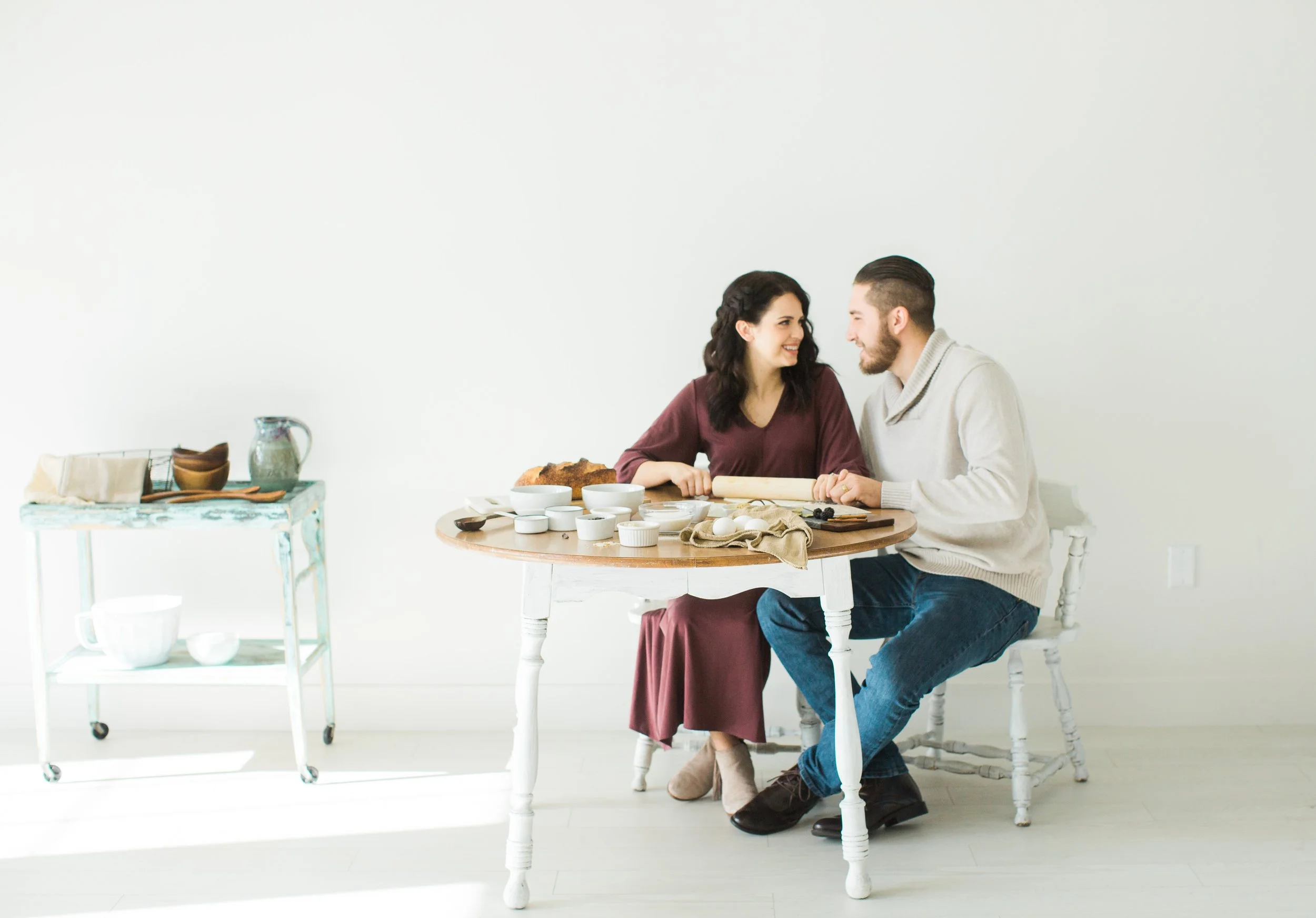 A woman and man sitting at a white table, smiling at each other. The table has baking ingredients, eggs, and a rolling pin. The setting is bright with a plain white wall background.