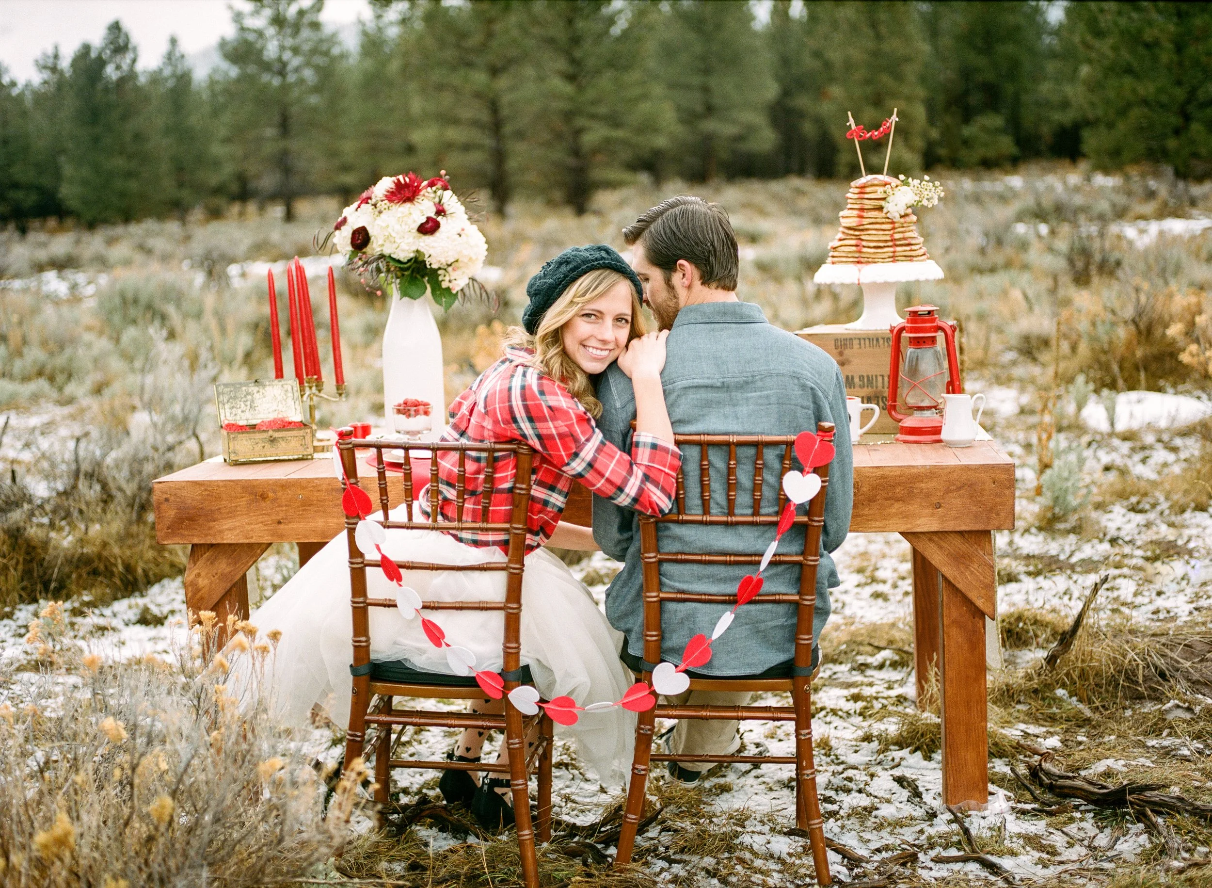 A couple sitting at a wooden outdoor table in a natural setting with trees and grass, decorated with flowers, hearts, and pancakes with syrup, celebrating an event.