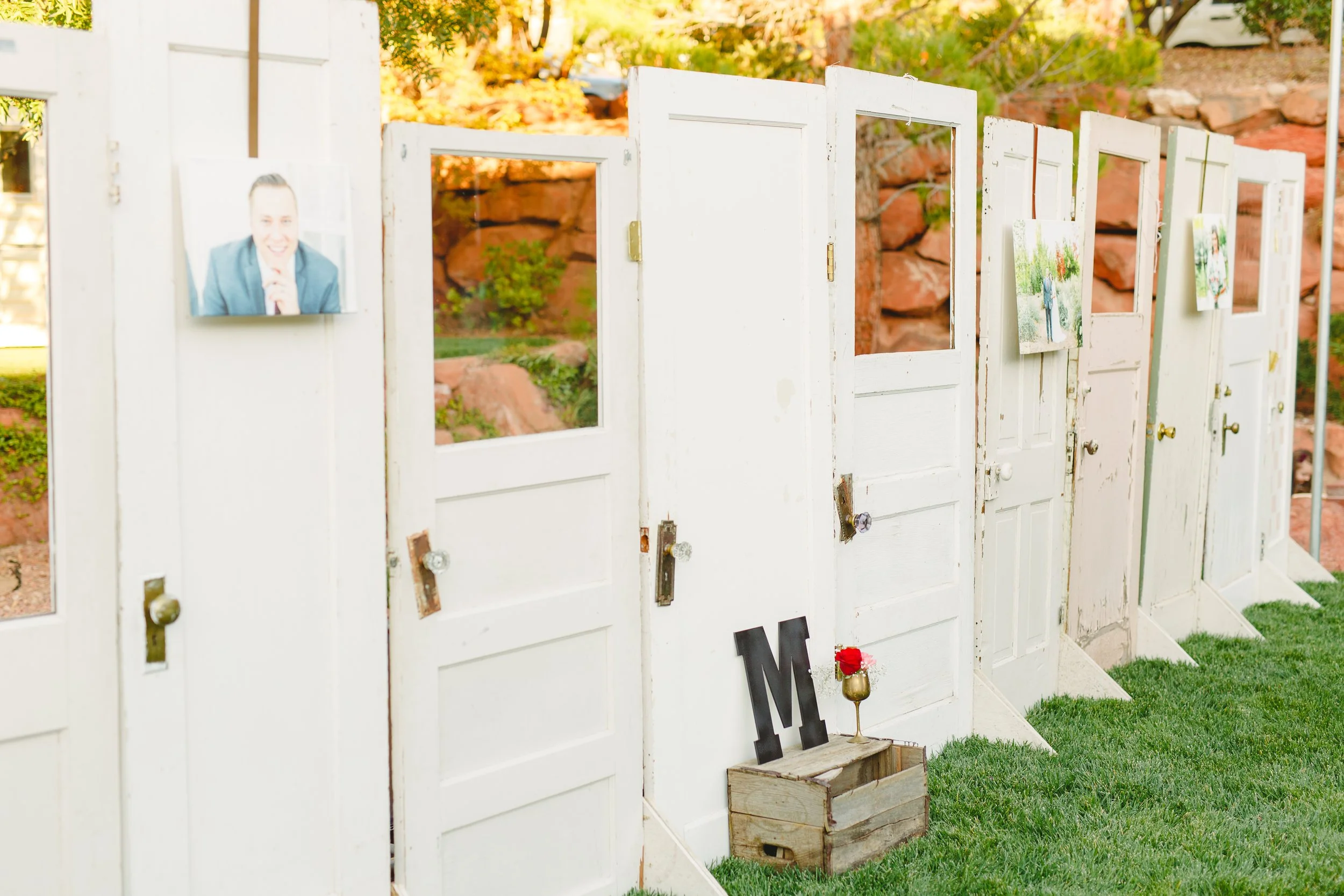 White wooden memorial display with open doors, featuring photographs, candles, and a black letter 'M' on a small wooden crate, set outdoors on green grass with autumn foliage in the background.