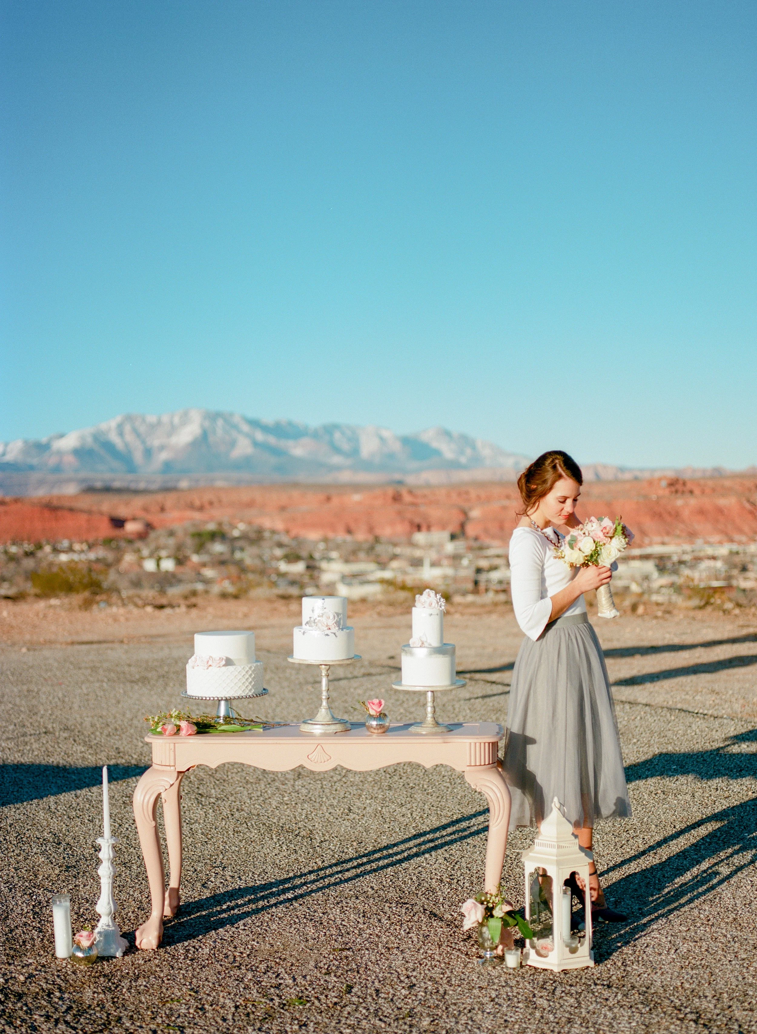A woman in a gray skirt and white top standing outdoors at a wedding or celebration, holding a bouquet of flowers. A pink decorated table displays white cakes and flowers, with candles and lanterns around it. The background features mountains and a clear blue sky.