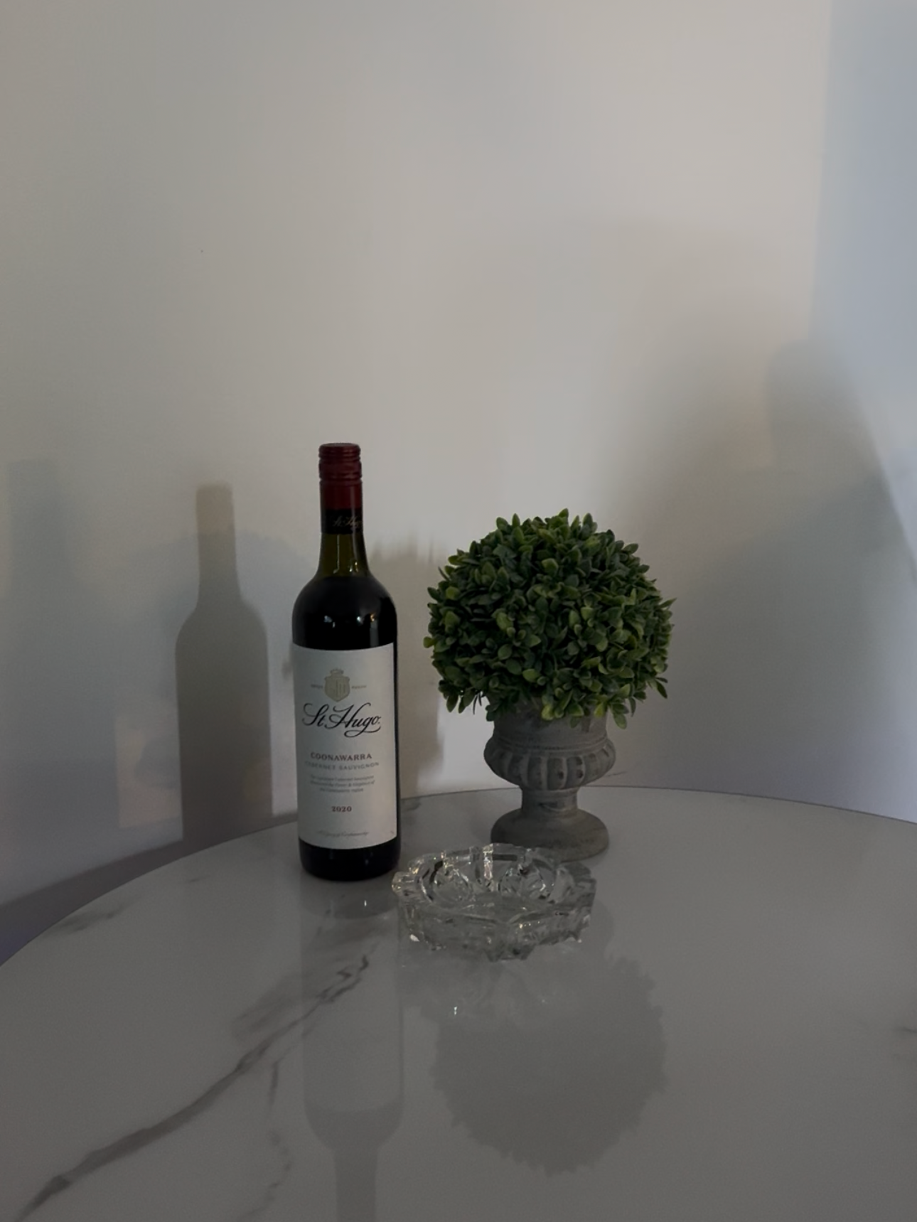 A bottle of red wine, a potted green plant in a decorative gray planter, and an empty glass ashtray on a white marble table against a white wall.