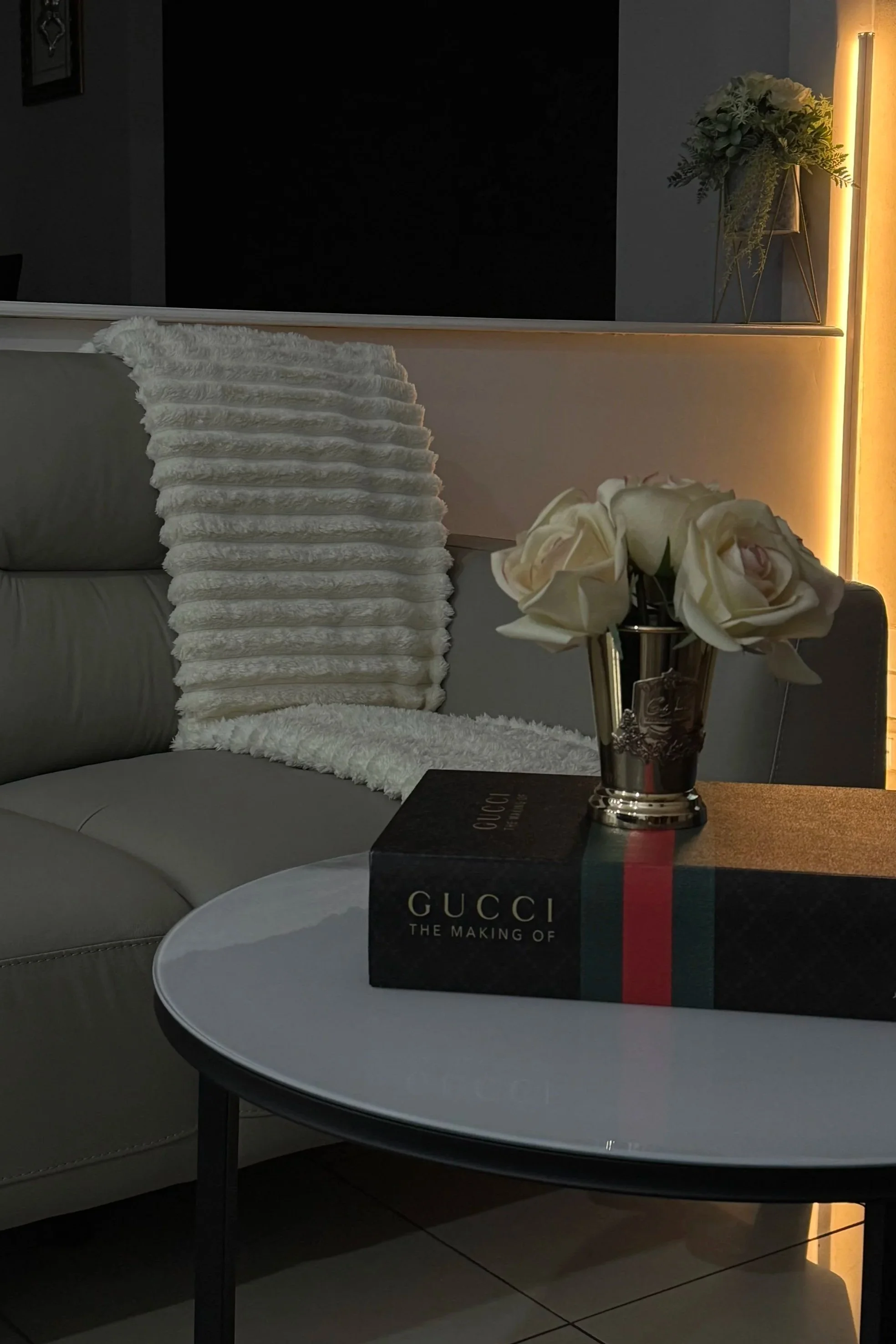 A living room corner with a beige sofa, white fur blanket, black and Gucci book on a round white table, and a gold vase with white roses, illuminated by warm lighting.
