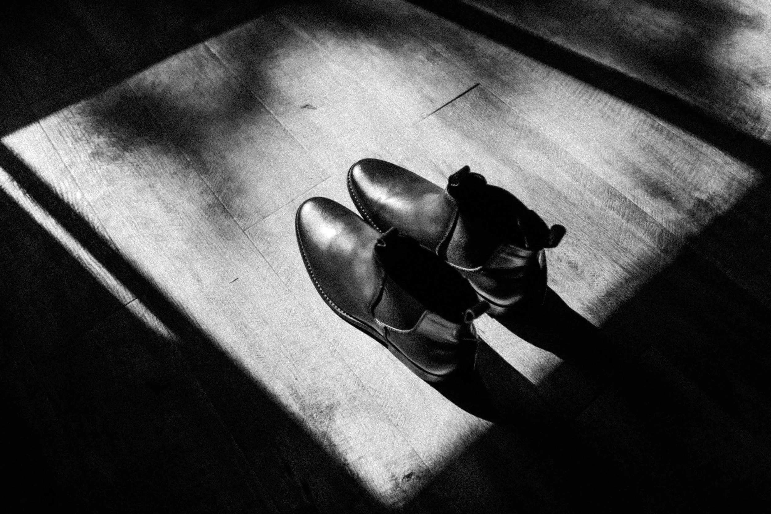 Black and white photo of shiny shoes on a person's feet, standing on a wooden floor near a doorway.