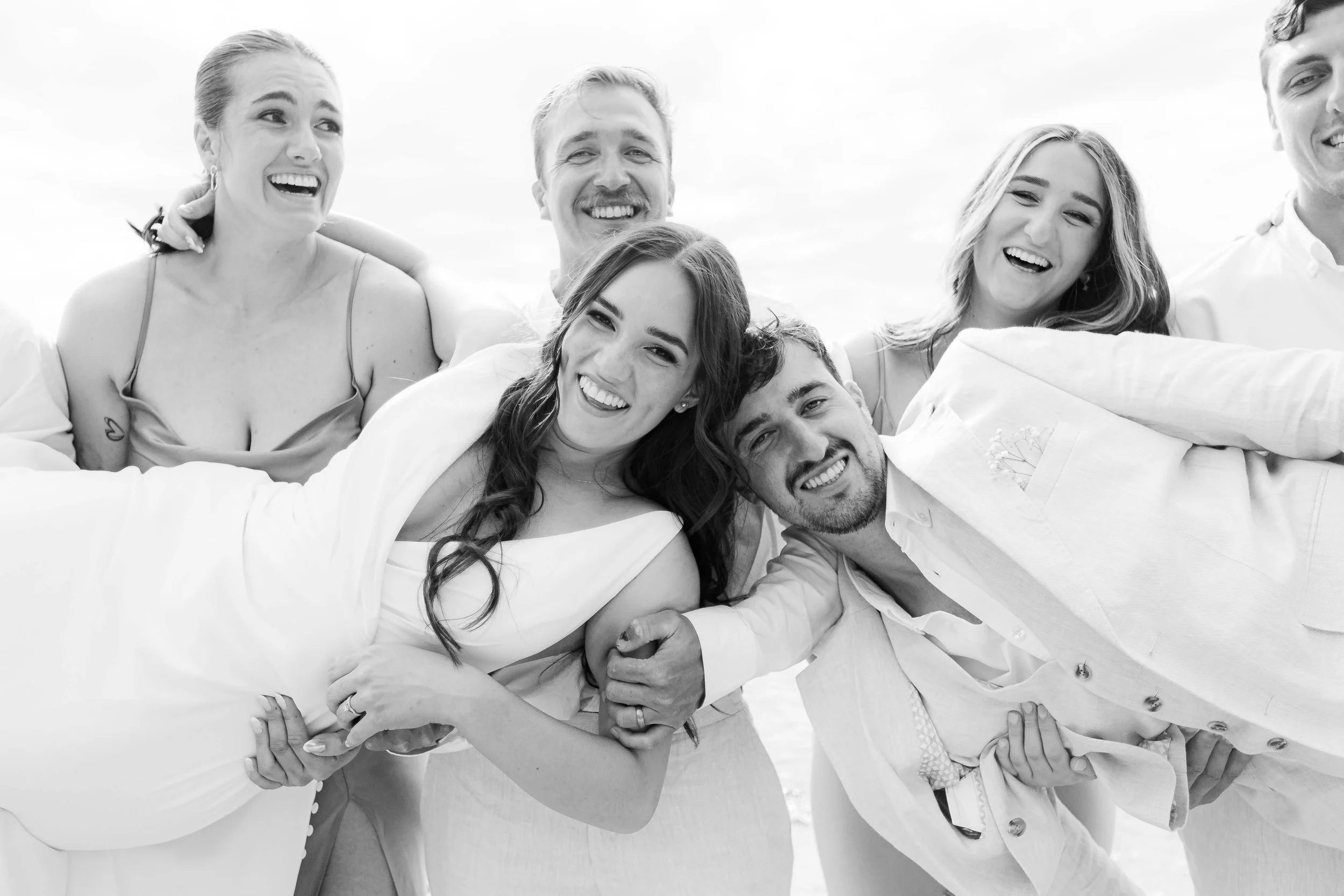 Group of six happy young adults, four women and two men, playing and laughing outdoors, with three women in front and three men behind them, under a partly cloudy sky.