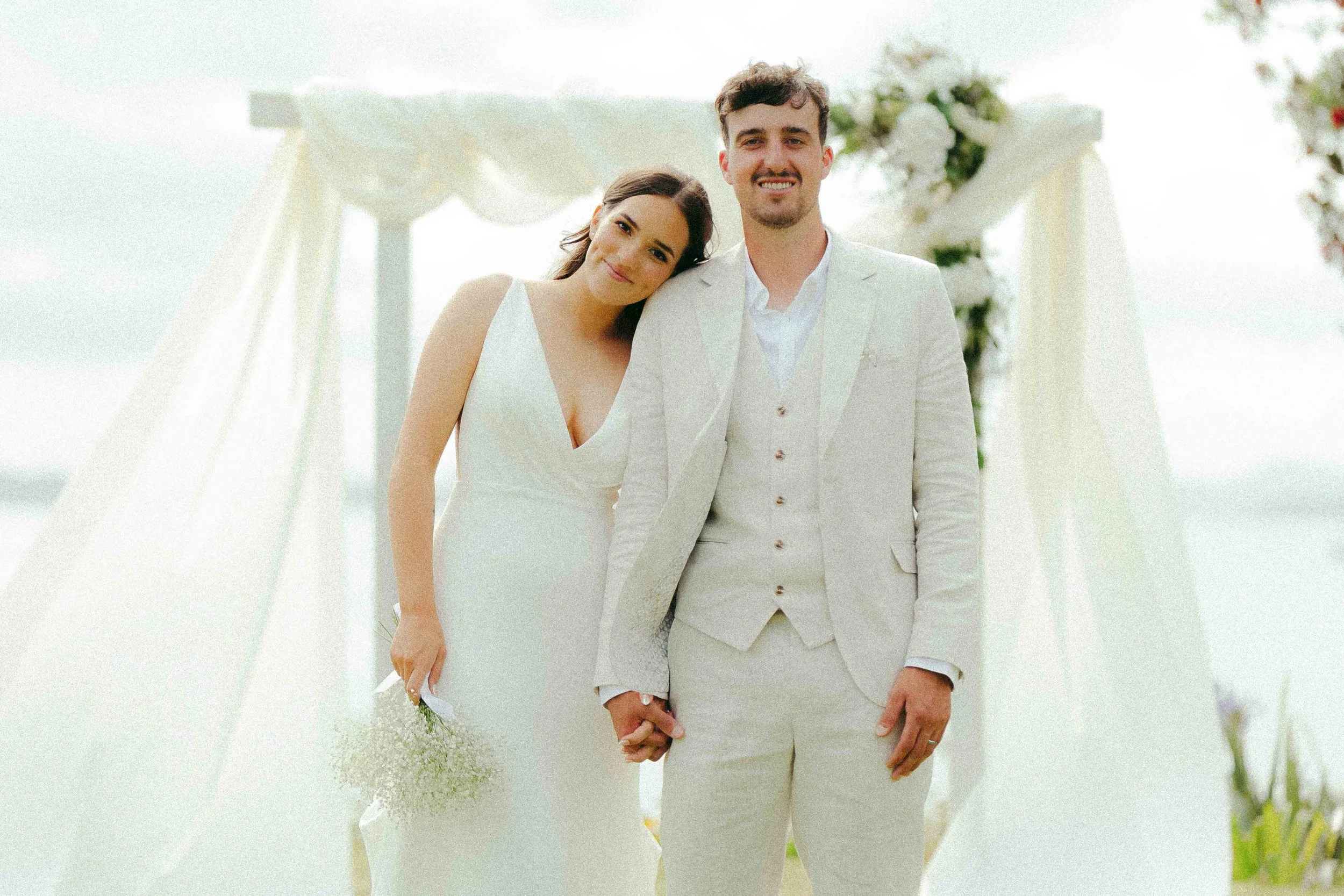 A bride and groom holding hands during their outdoor wedding ceremony, standing under a white floral arch.