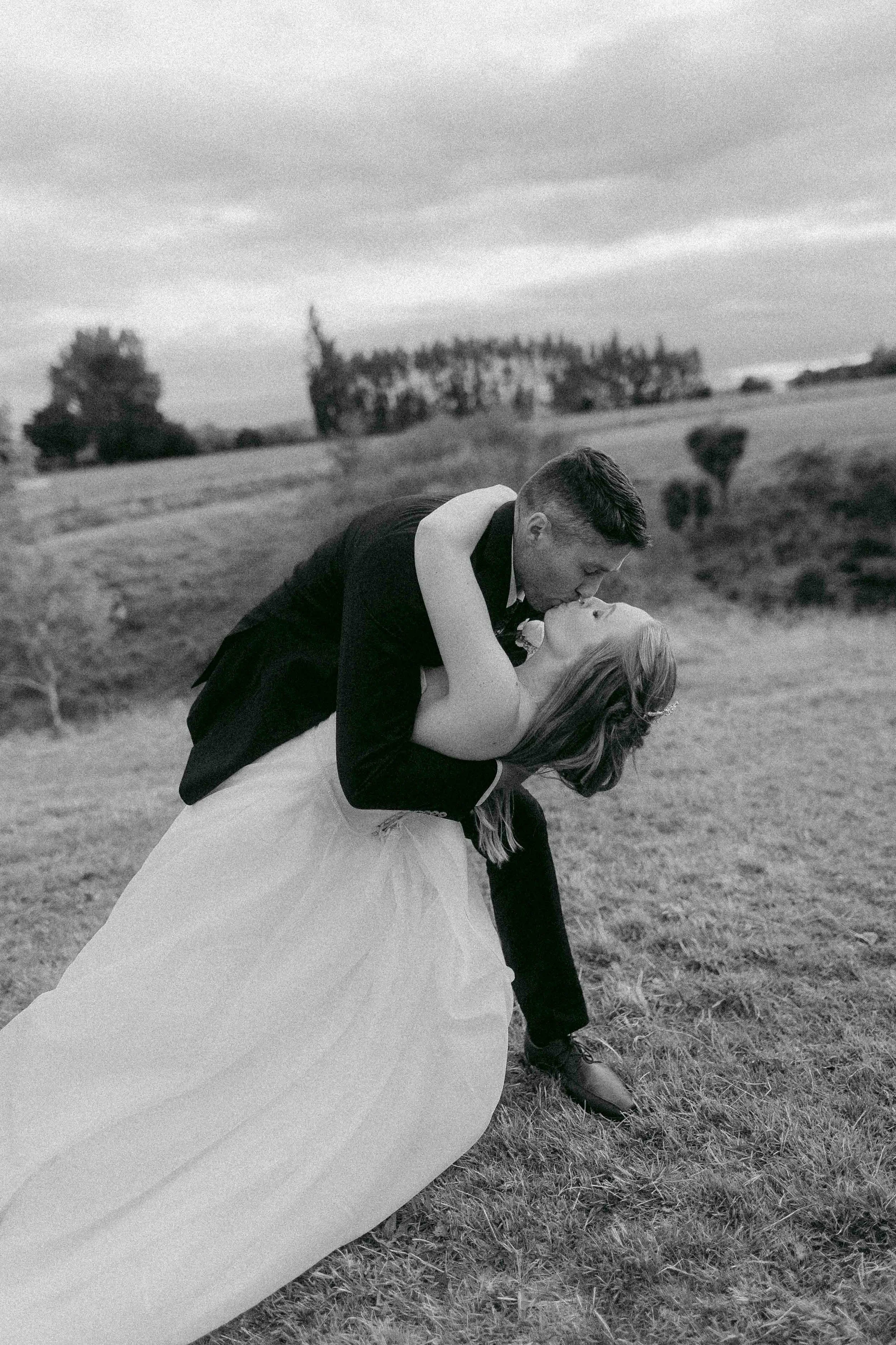 A black-and-white photo of a couple kissing outdoors in a grassy field, with trees in the background and cloudy sky overhead.