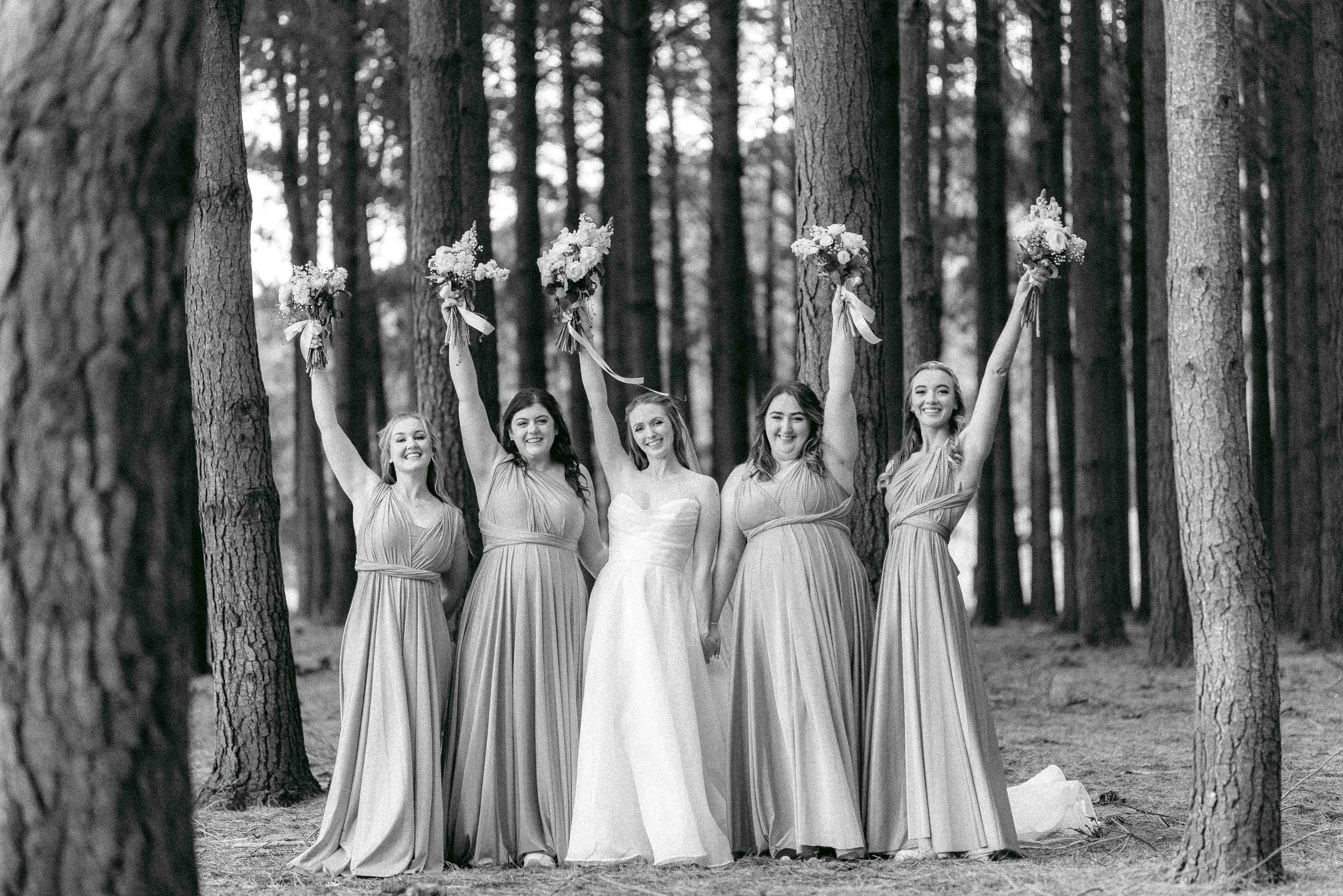 A group of five women dressed in long gowns, standing in a forest with tall trees. The women are smiling and holding bouquets of flowers raised in the air.