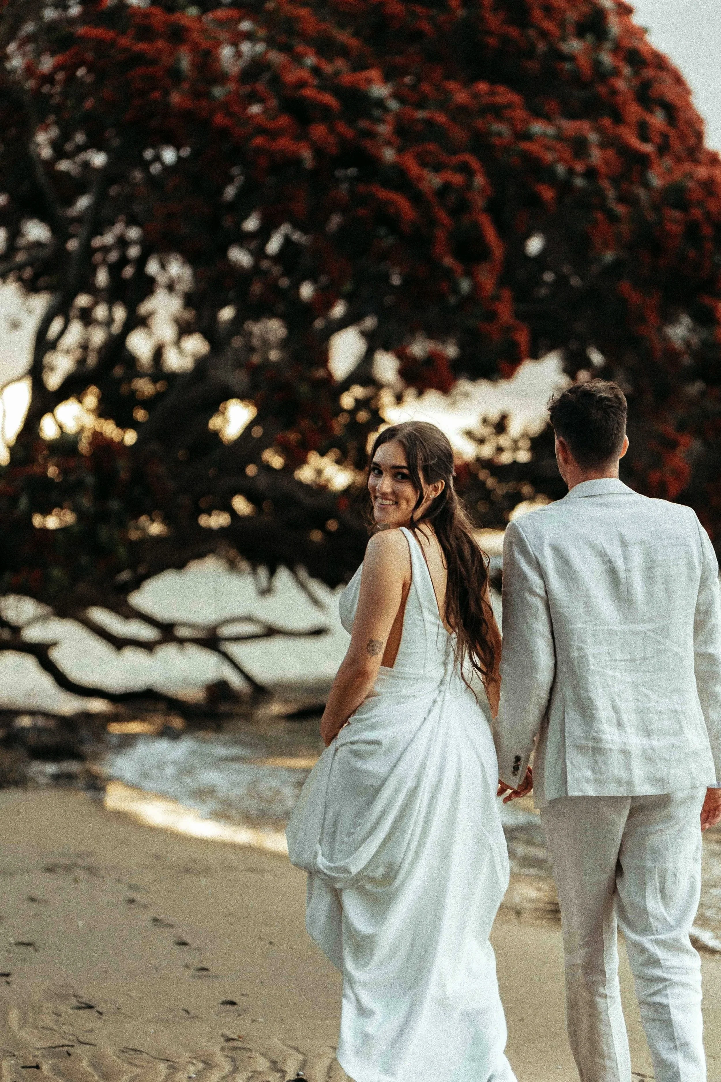 A newlywed couple holding hands and walking along the beach at sunset, with a large tree with red leaves in the background.