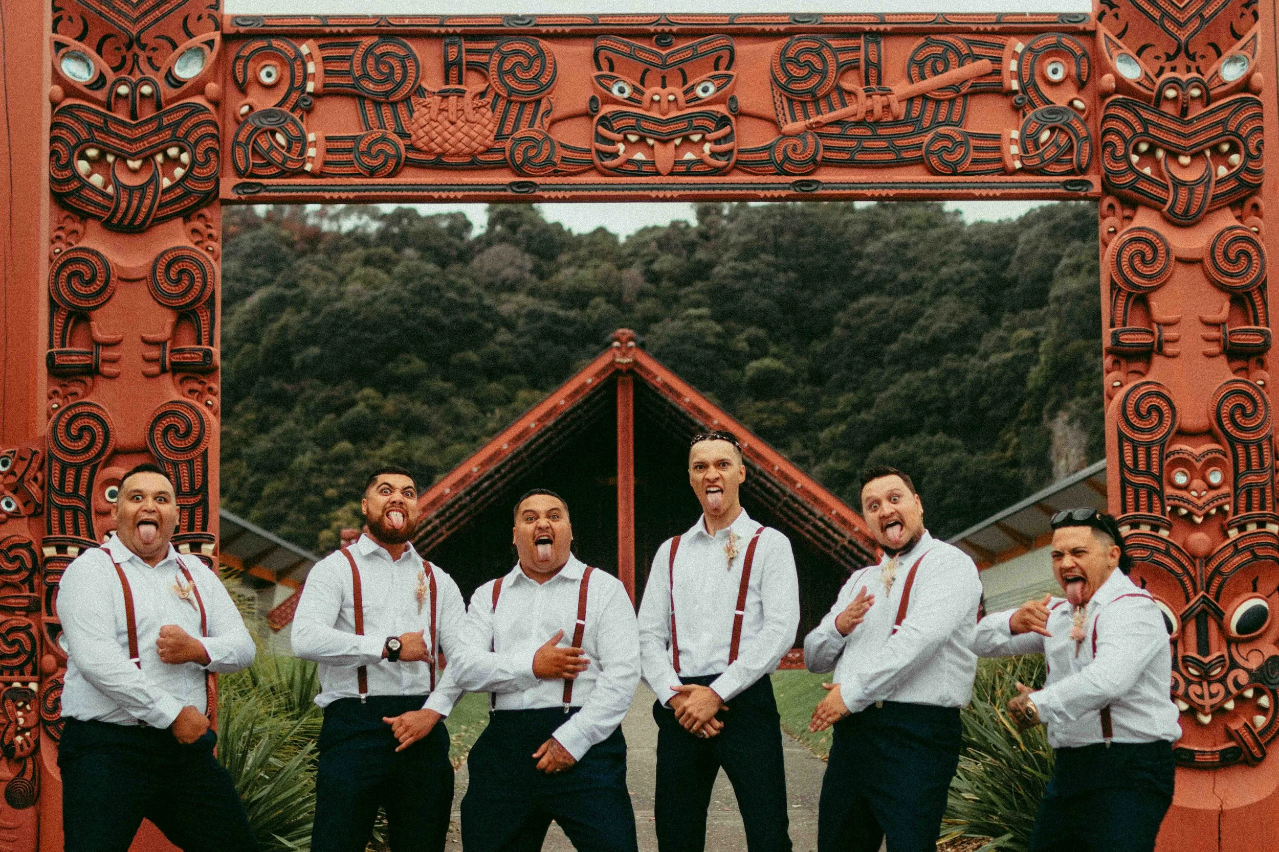Group of men wearing white shirts with suspenders posing with tongues out in front of a carved Maori-style gate with greenery and hills in the background.