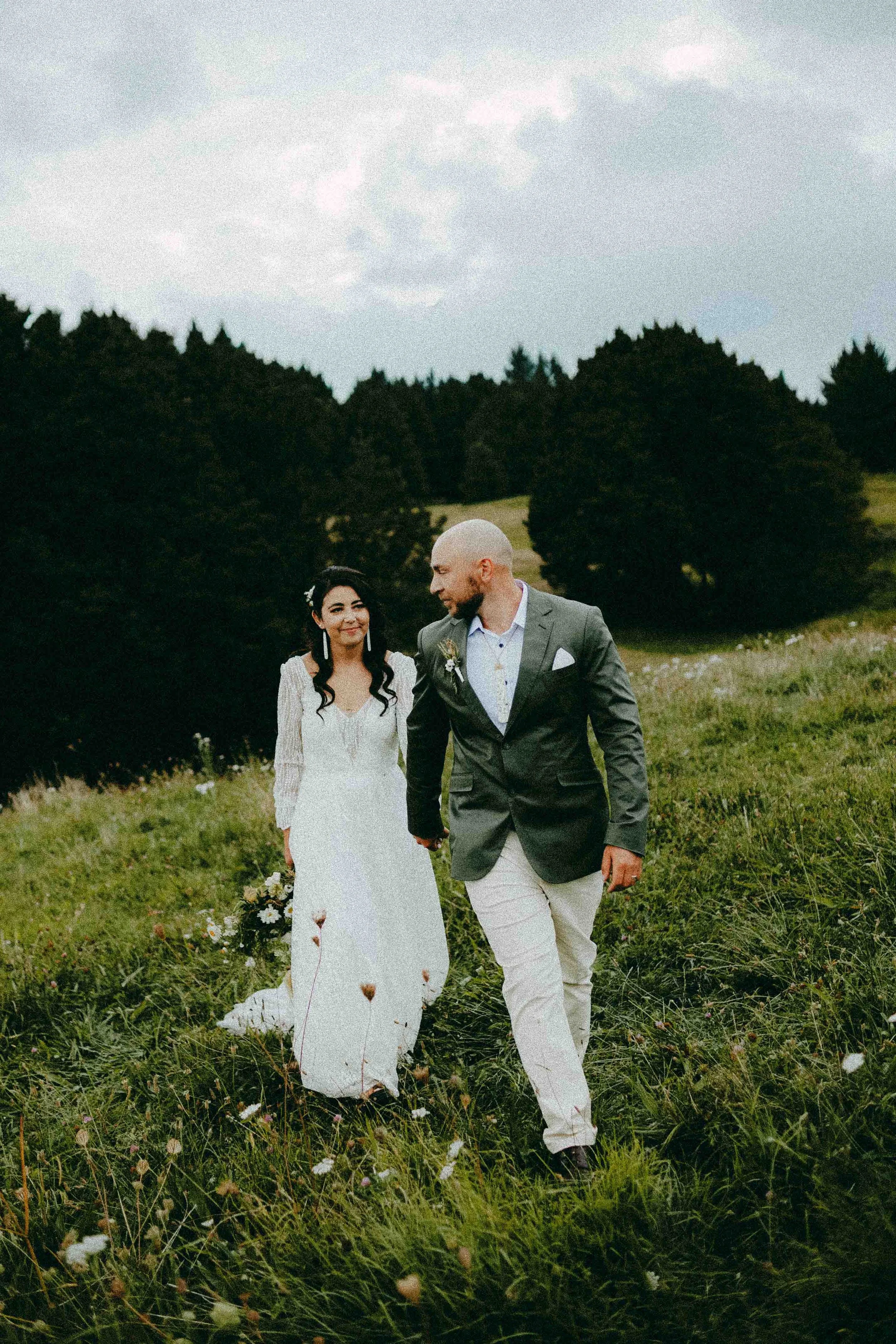 A bride and groom walking hand in hand through a grassy field with wildflowers, trees and cloudy sky in the background.