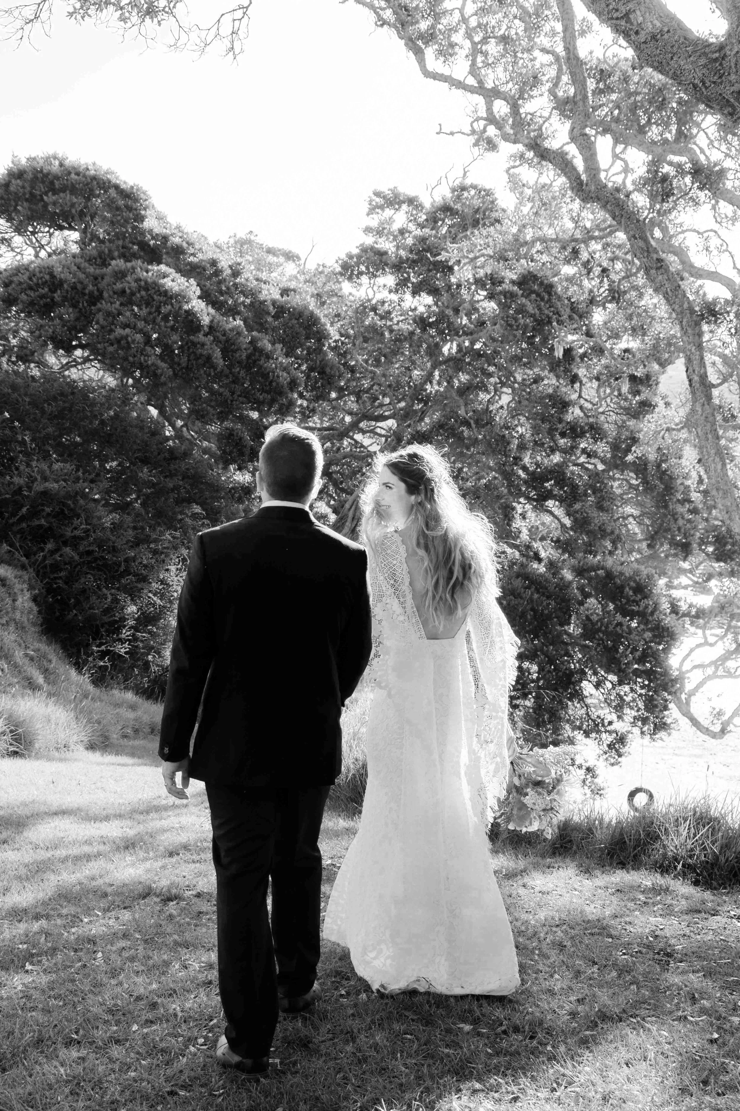 A black-and-white photo of a bride and groom walking outdoors through tall trees. The bride is wearing a long lace wedding gown and holding a bouquet, and the groom is dressed in a suit. They are walking away from the camera, and the sunlight filters through the trees.
