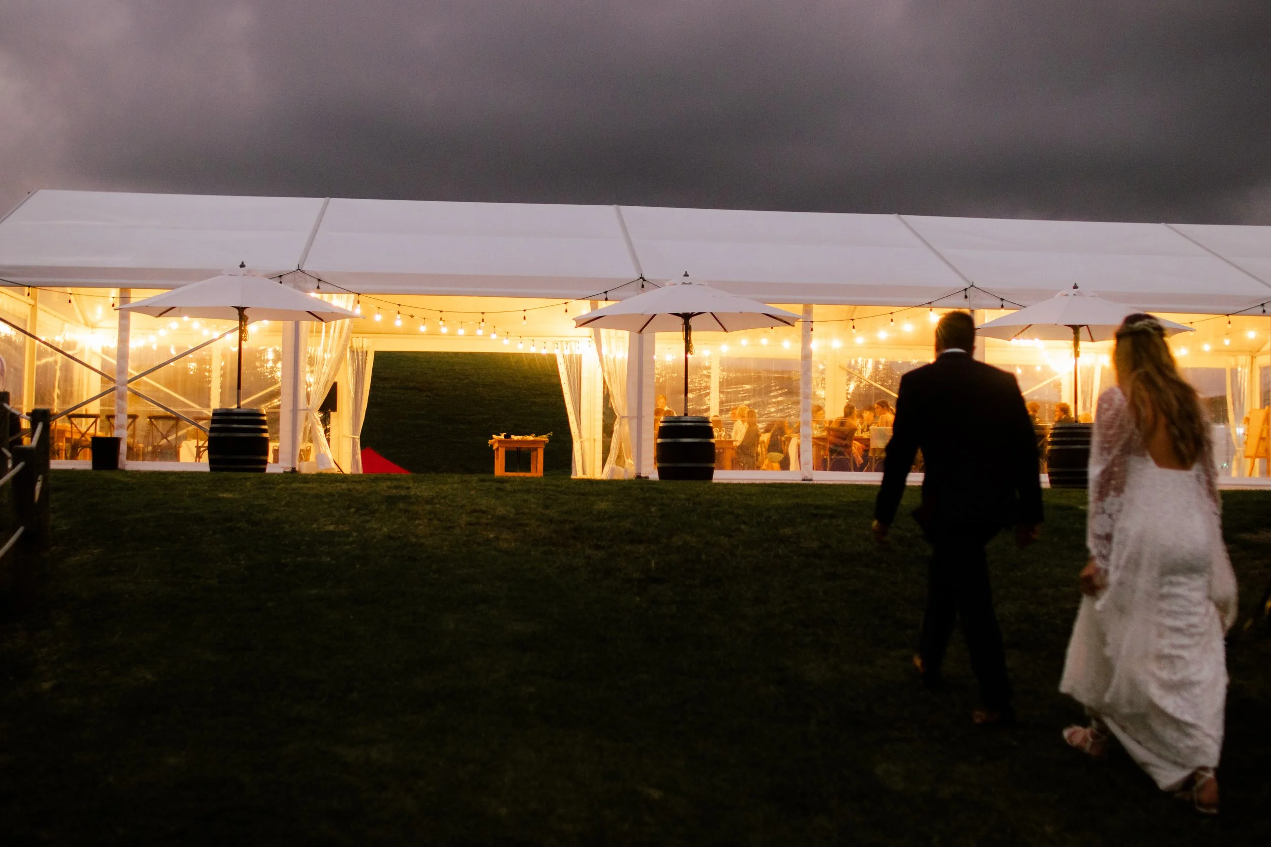 People walking on grass toward a well-lit event tent with string lights and umbrellas in the evening, dark cloudy sky above.