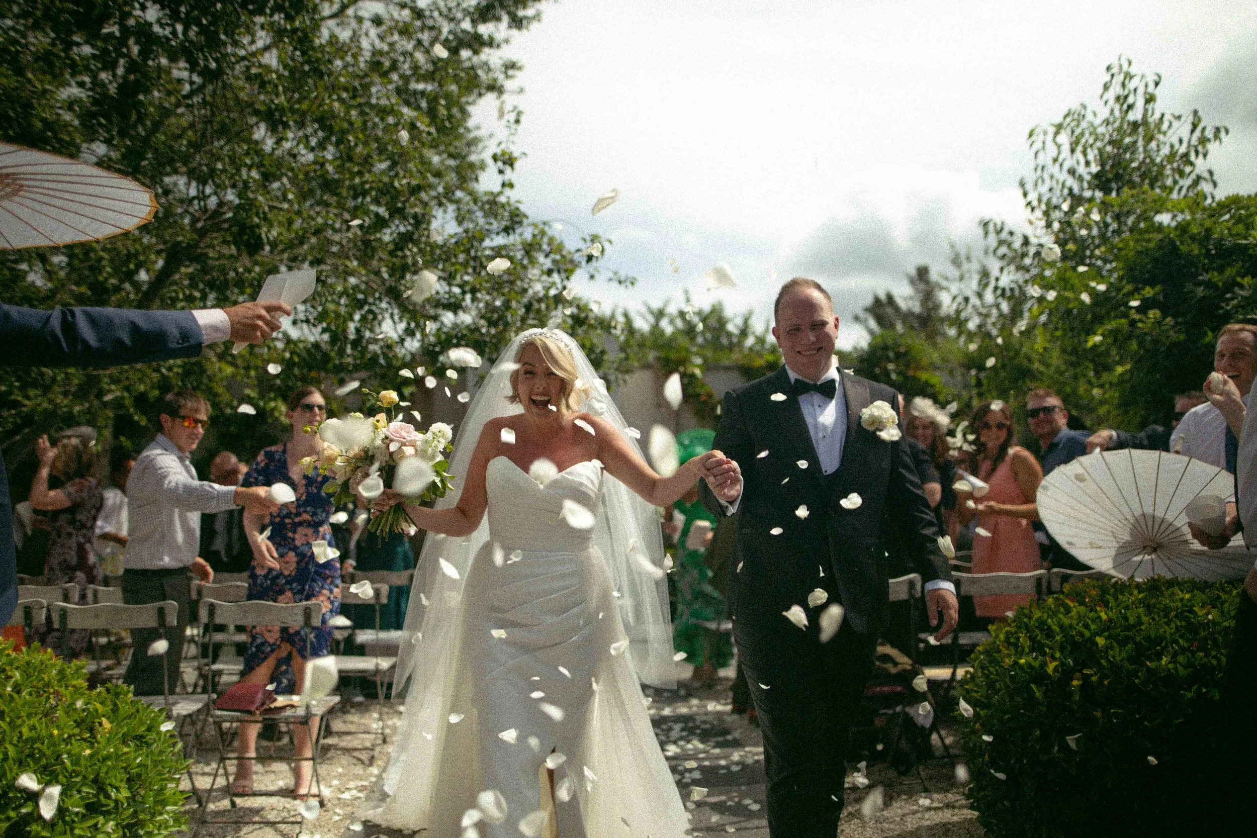 Bride and groom walking outdoors at a wedding ceremony, surrounded by guests throwing flower petals. The bride is wearing a white wedding dress and veil, holding a bouquet, and the groom is dressed in a black tuxedo with a white shirt and bow tie. There are trees and chairs with guests in the background.