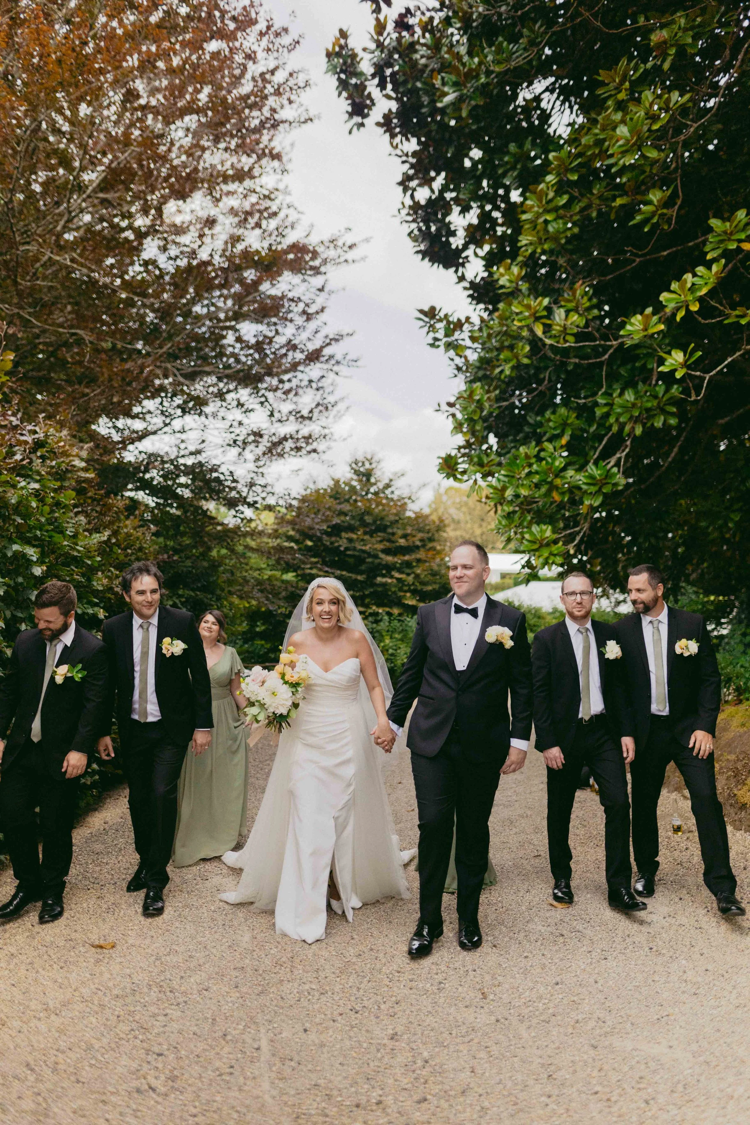 A group of people, including a bride and groom, walking outdoors on a gravel path surrounded by trees during a wedding celebration.